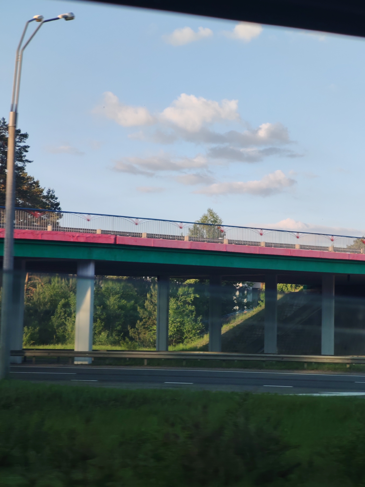 The image depicts a bridge over a highway. The bridge is supported by white pillars and painted with a green and red color scheme. It has a silver railing with red accents. There are trees visible behind the bridge supports. The highway is visible with white lane markings and a guardrail. Grass and vegetation are visible in the foreground. A lamppost is present on the left side of the image. The sky above is blue with scattered white clouds. The location is unknown.