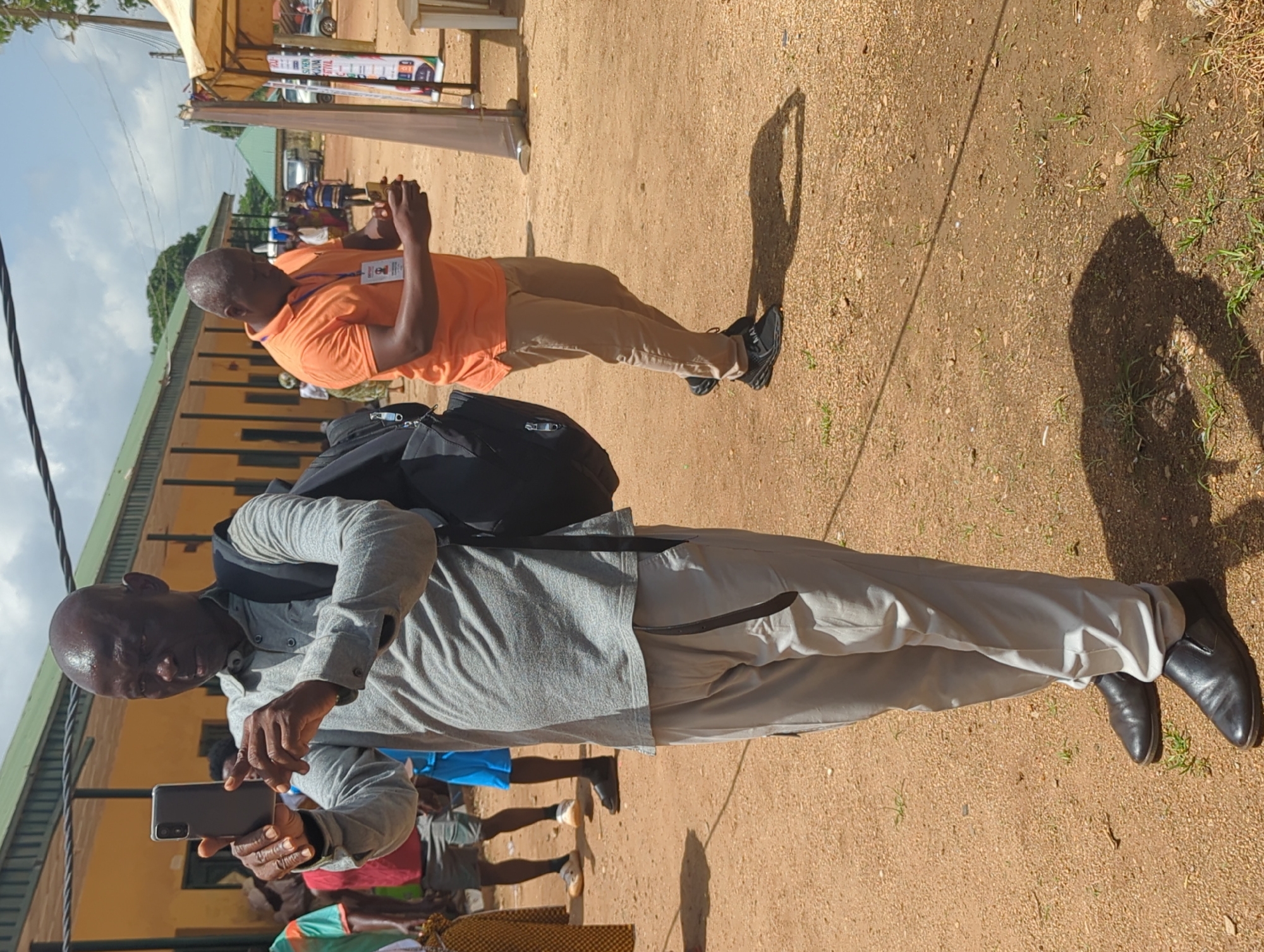 The image shows two men standing on a dirt ground in Maigizo, Nigeria. One man is wearing a grey shirt, light grey pants, and black shoes. He has a black backpack and is holding a black smartphone, seemingly taking a picture. The other man is wearing an orange shirt, beige pants, and black sneakers. He is also holding a smartphone and appearing to take a picture. The background includes a building with a green roof and yellow walls. Other people are visible in the background. The lighting suggests it is daytime. A temporary tent structure with signs is present. The ground is primarily dirt with sparse vegetation.
