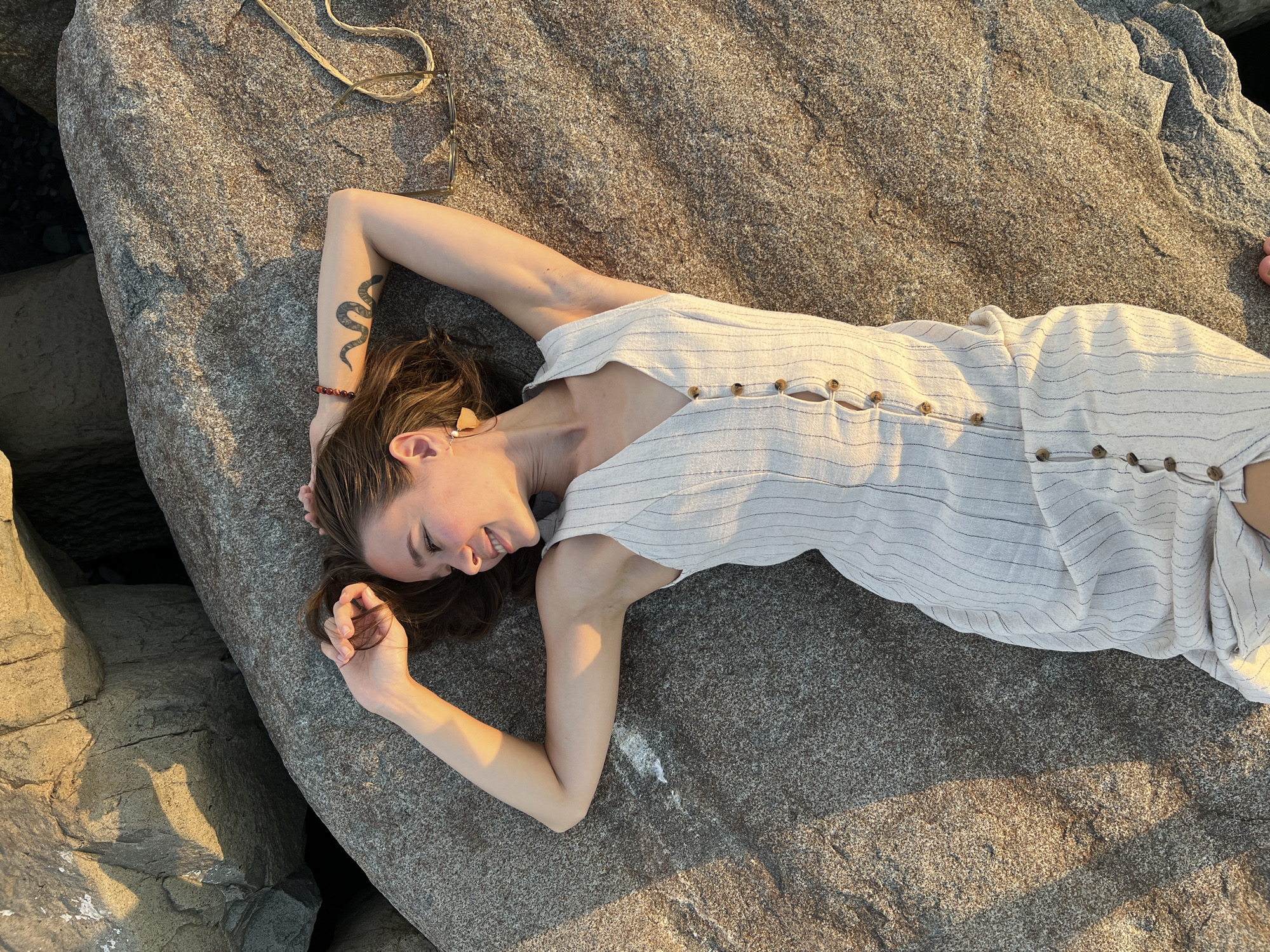 A young woman with brown hair, wearing a light-colored striped dress and earrings, lies on her back on a large rock formation. Her arms are raised above her head, and she is looking to her left with a smile. She has a snake tattoo on her left arm and a beaded bracelet. Sunglasses rest on the rock near her right arm. The scene is bathed in the warm light of sunset, casting shadows on the textured surface of the rocks. The location appears to be a rocky coastline, potentially near Batumi, Georgia, given the natural rock formations and oceanic scenery.
