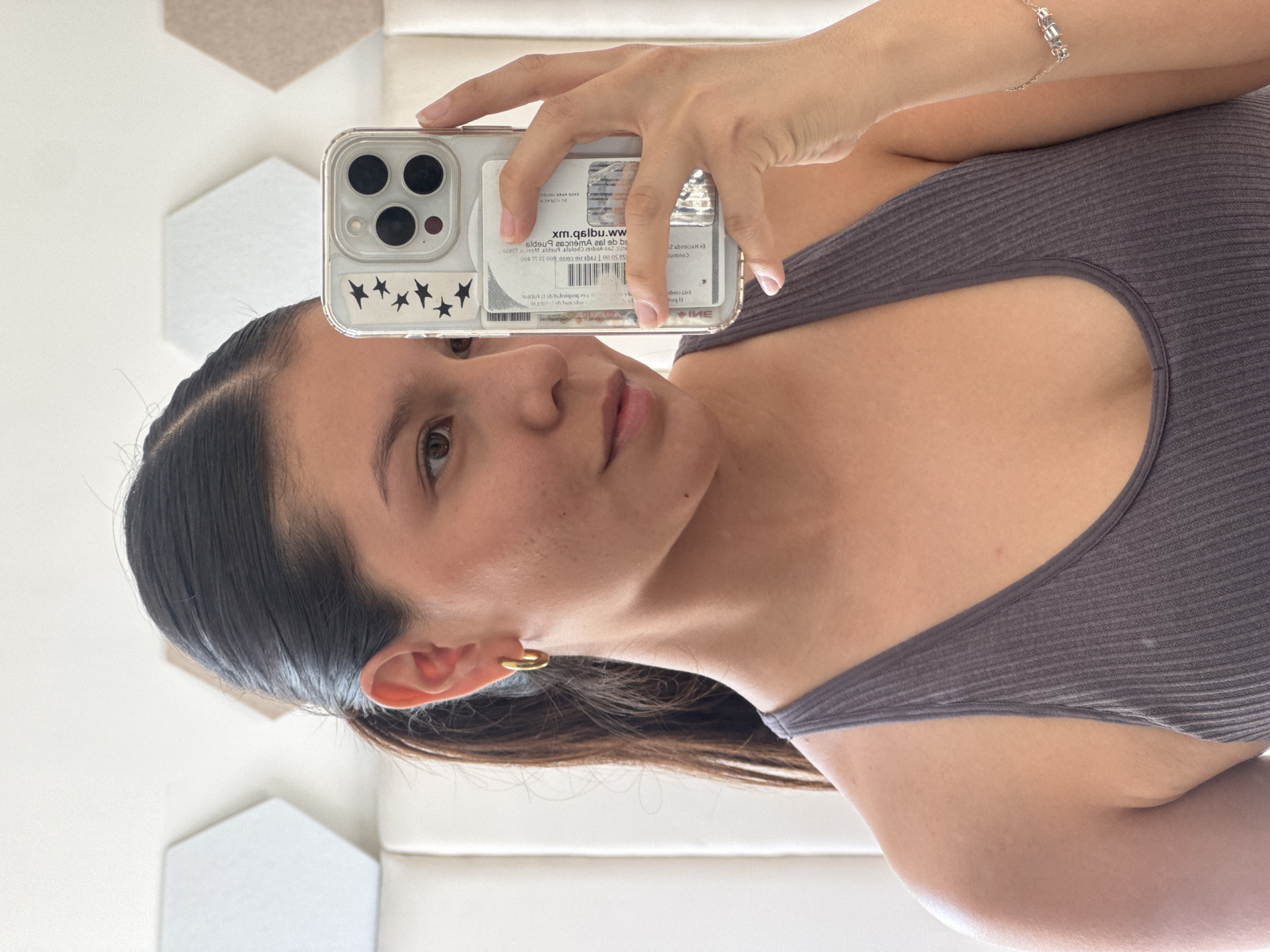 A woman is taking a selfie with her phone. The phone has a clear case with star stickers and a card inserted in the back. The woman has dark hair pulled back, a gold hoop earring, and a grey top. She is wearing a bracelet on her right wrist. The background includes geometric shapes and a white wall. The card visible in her phone case contains the text "xm.qslbu.ww" and other printed details in Spanish. Based on the partial text of "San Francisco Ocotlán" on the card visible in the phone case, the card may be related to this location in Ocotlán, Mexico.