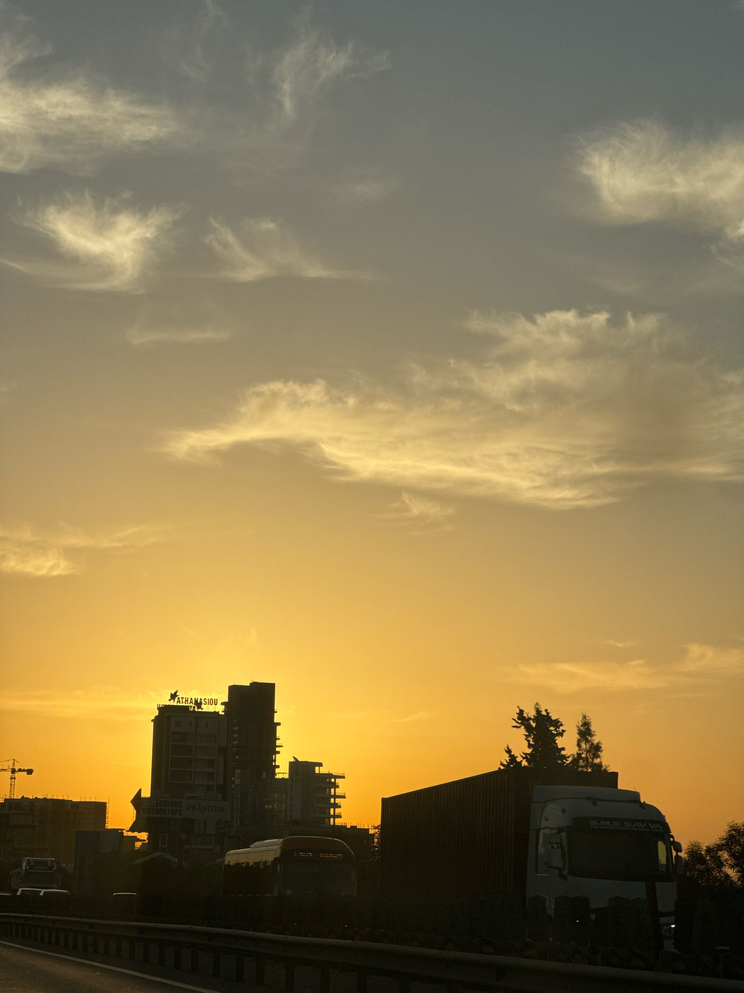 The image captures a silhouetted view of a highway and buildings against a vibrant sunset in Mouttagiaka Community, Cyprus. In the foreground, a highway is visible with a concrete barrier running along the side. Two lanes of traffic are suggested by the road markings, though no vehicles are actively moving in the immediate foreground.
Dominating the mid-ground are the silhouetted forms of several buildings. A prominent multi-story structure, likely an office building or hotel, stands tall with signage that reads "ATHANASIOU" and "COLOURS PRASINOS KITCHEN TOPS". To the left of this building, a construction crane is partially visible, indicating ongoing development in the area. Further to the right, a large articulated truck with a refrigerated trailer is parked on the side of the road, its front cab illuminated by headlights. Several trees are silhouetted behind the truck.
The sky is a striking gradient of orange and yellow, transitioning to a muted grey at the top, filled with wispy clouds illuminated by the setting sun. The overall impression is one of urban development meeting the natural beauty of a sunset.