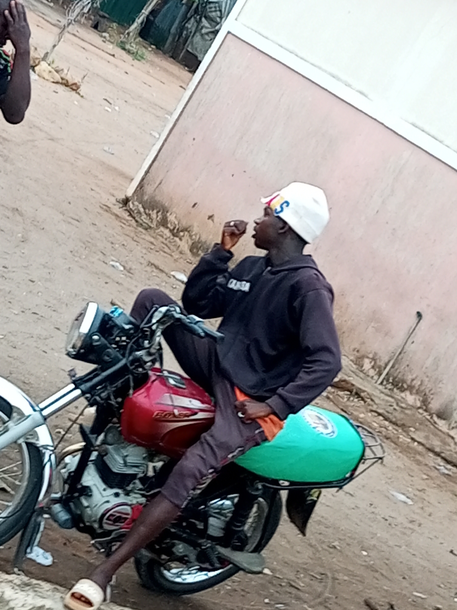 In Tumfure, Nigeria, a young Black male is sitting on a red motorcycle. He is wearing a white beanie with colorful letters on it, a black hoodie, and dark pants. The motorcycle has a green bag on the back. The background consists of a sandy ground and a light-colored building. The image is taken from a low angle, looking up at the male and the motorcycle.