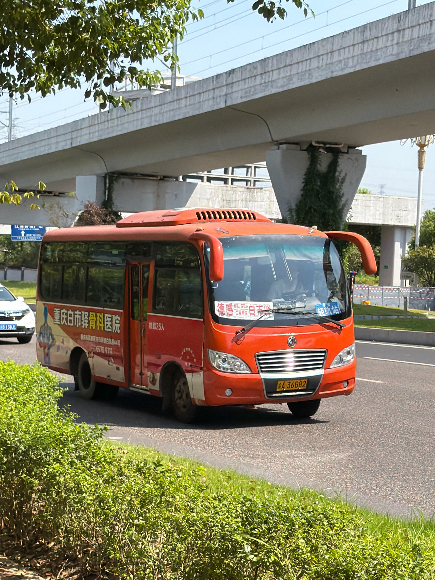 A bright orange bus, identified by the license plate "A 36682," is driving on a paved road. The bus features advertisements on its side, one of which displays Chinese text including "重庆市白市骨科医院" (Chongqing Baishi Orthopedic Hospital) and a phone number. On the front of the bus, a blue sign with Chinese characters indicates "轻轨站" (Light Rail Station). The bus has a passenger inside, visible through the windshield. In the background, an elevated concrete structure, likely an overpass or bridge, looms over the scene. Green foliage lines the side of the road in the foreground. The sky is clear and blue, suggesting a sunny day. The scene appears to be in the city of Jiangjin, China, given the text on the bus.