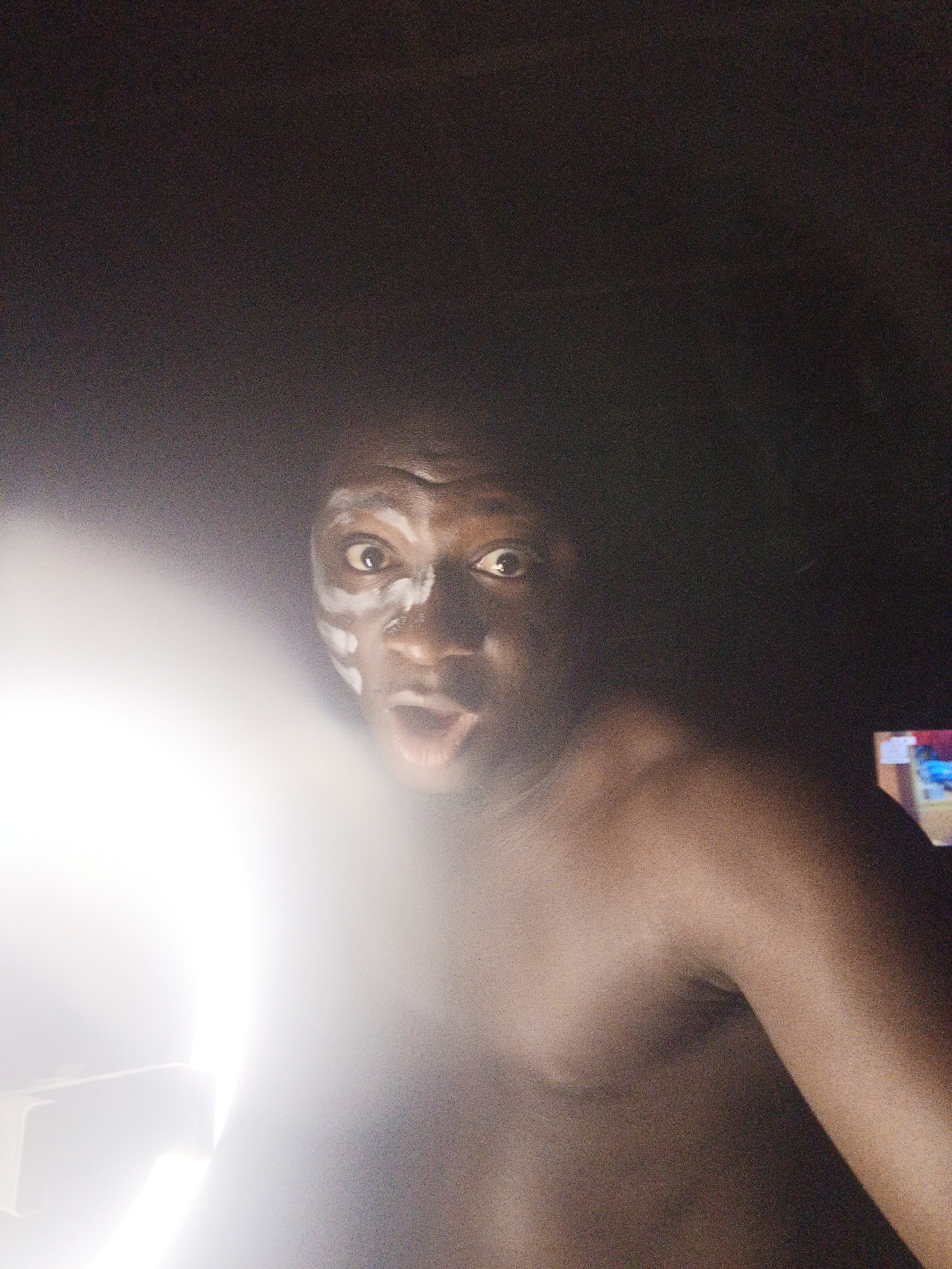 An adult individual with dark skin is prominently featured, looking directly at the viewer with a wide-eyed and open-mouthed expression, conveying surprise or shock. White markings, possibly face paint, are visible on their forehead, around their eyes, and on their cheeks. The individual appears shirtless, with their bare shoulders and chest visible.
A very bright, circular light source is positioned on the left side of the frame, creating a strong lens flare that obscures the lower-left portion of the image and casts dramatic lighting on the subject's face and body. The rest of the scene is dark, suggesting a low-light environment, possibly indoors. In the dimly lit background to the far right, a rectangular object with colorful elements, likely a television or computer screen, is faintly visible. The overall mood is intense and dramatic due to the expression and lighting. There is no visible text.