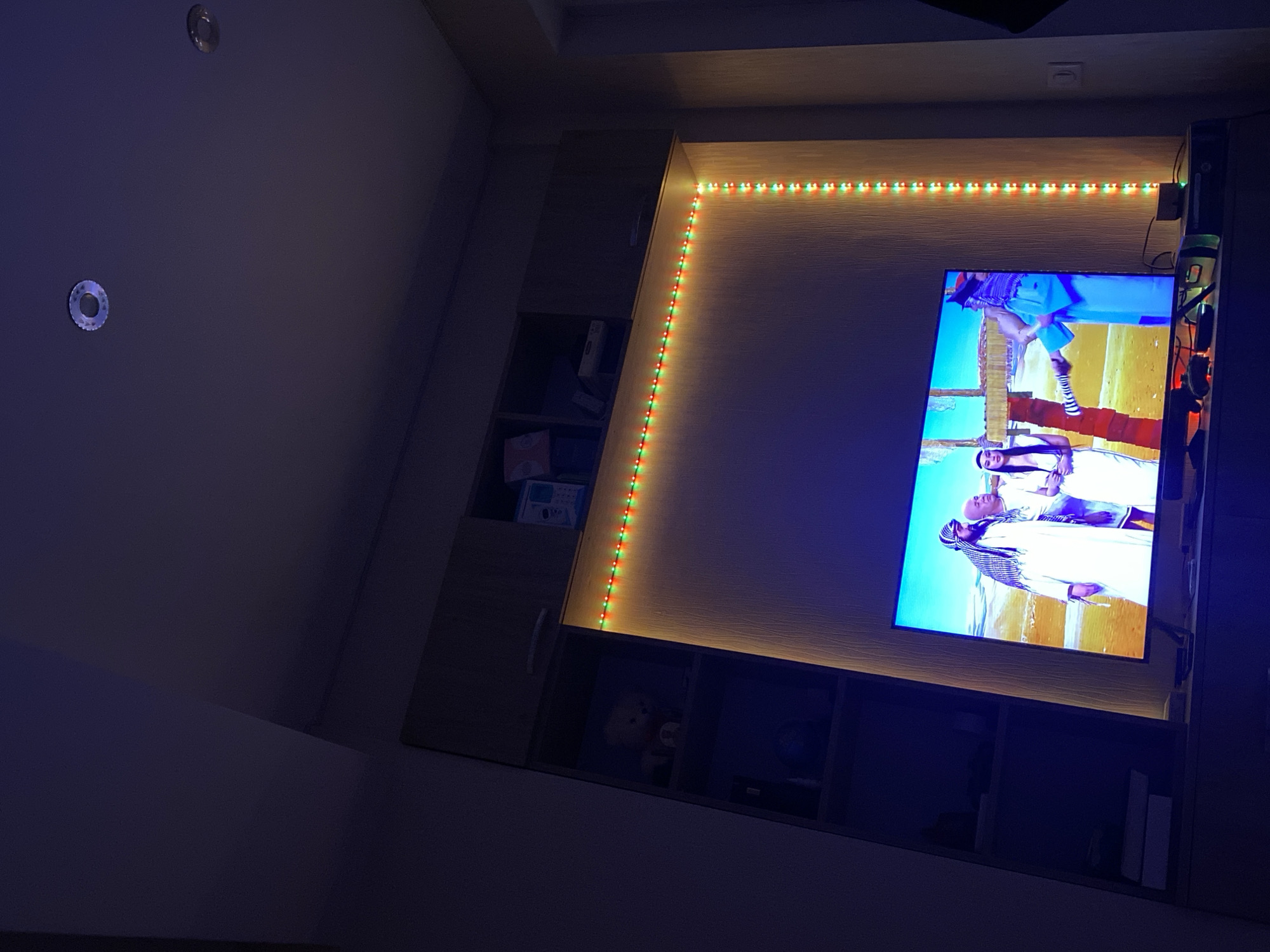 The image shows an indoor scene, likely a living room, with a large television screen displaying a scene featuring three individuals in traditional Middle Eastern attire. The television is mounted on a wall unit that has embedded LED strip lighting, alternating between red and green, framing the screen and running down the right side. To the left of the television are shelves within the wall unit, one of which contains a teddy bear. Above the wall unit, the ceiling features two circular recessed lights. The room appears to be dimly lit, with the primary illumination coming from the television and the LED strips. The overall impression is that of a contemporary entertainment setup within a residential space. There is no information available to determine the location within the city of Chișinău, Moldova.