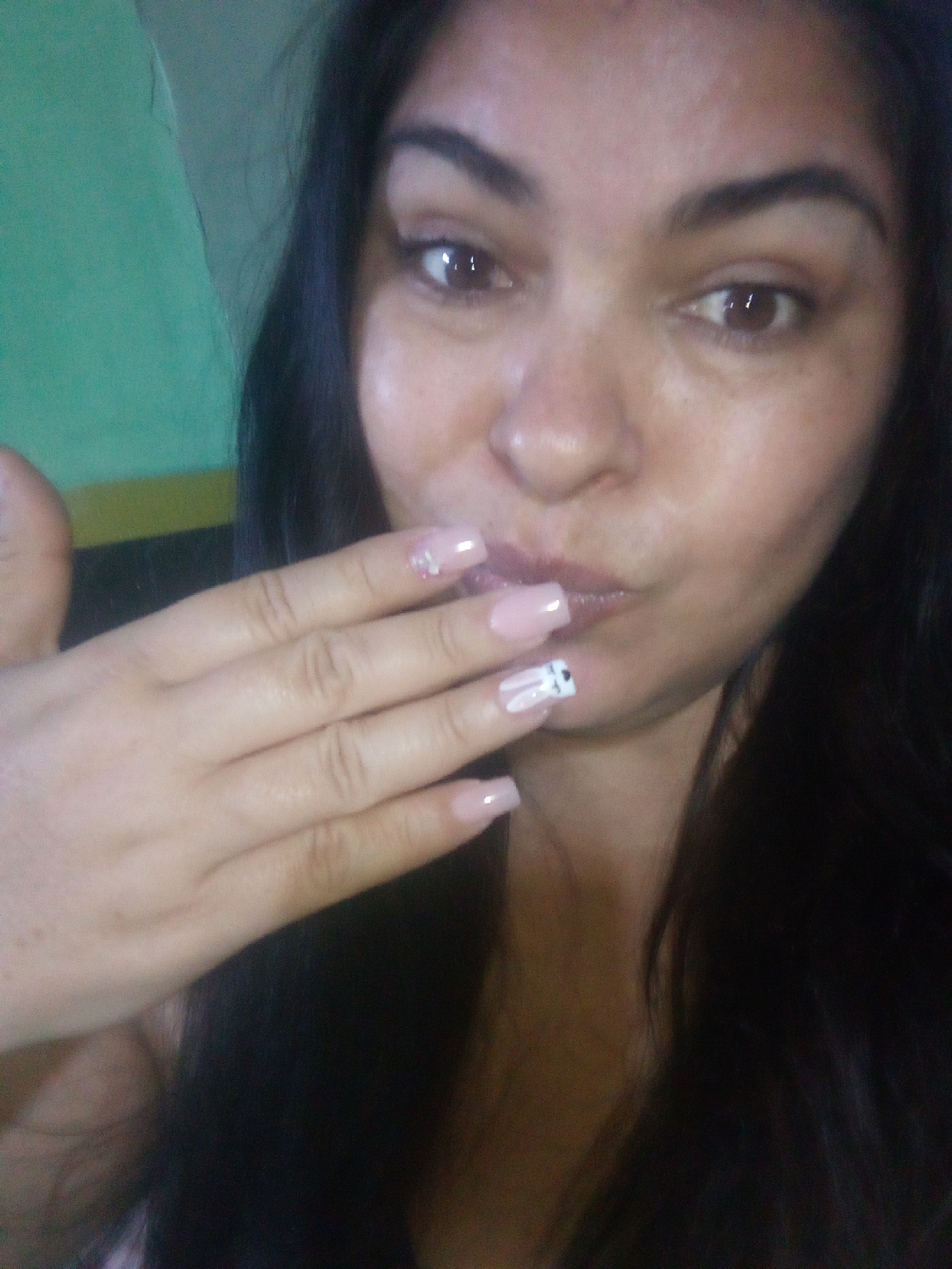 A woman with dark hair is posing for a selfie. She has fair skin and brown eyes. Her lips are pursed in a kissing gesture, and she is holding her hand up to her face, displaying her fingernails which are painted a light pink color with designs on two of them. The background is a mix of a green and yellow wall.