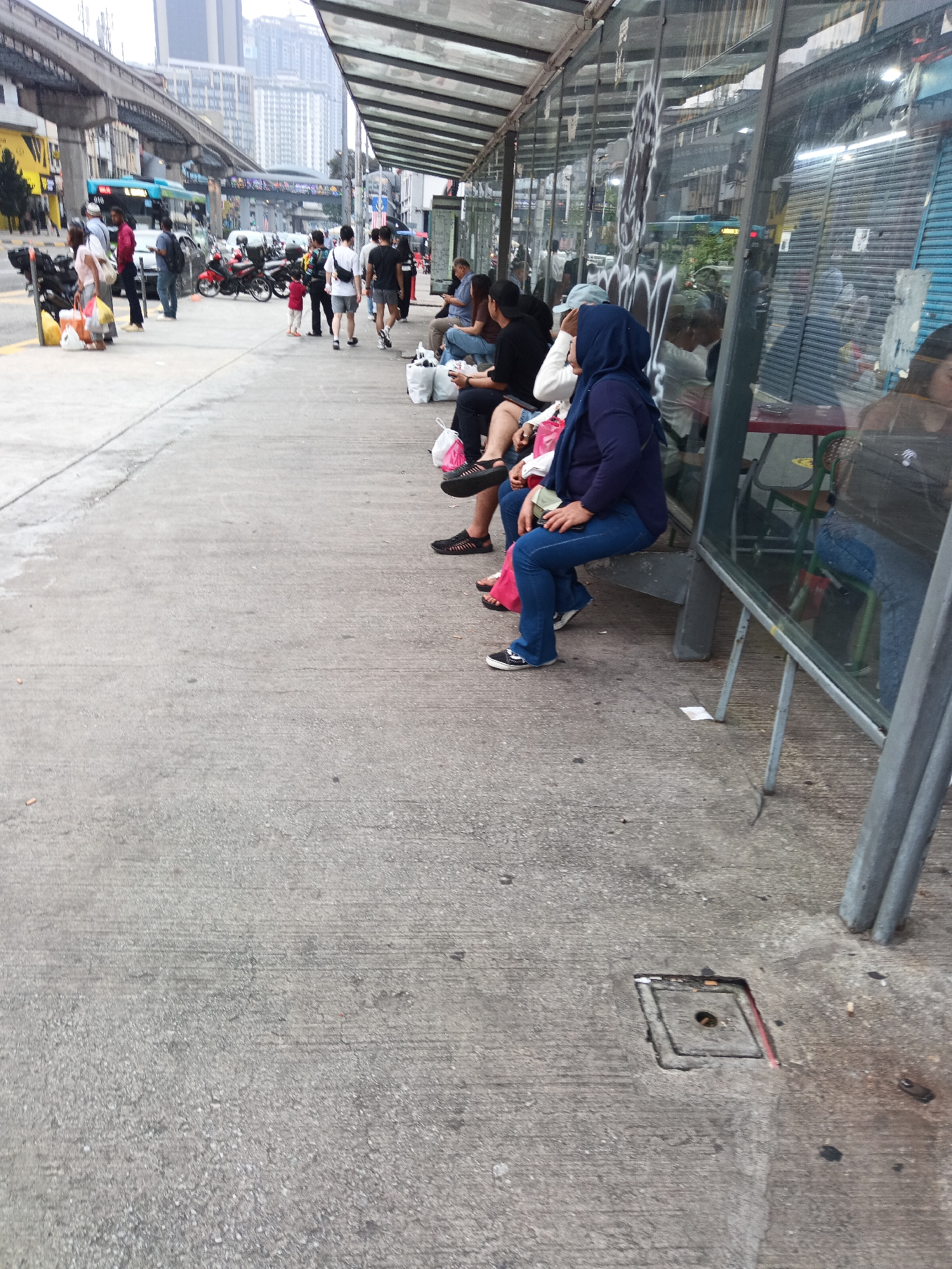 This image depicts a street scene in Kuala Lumpur, Malaysia, likely near a transportation hub, given the presence of a monorail track overhead. In the foreground, a concrete sidewalk is visible, with a drain cover embedded in it. Several people are seated at a bus stop shelter with a glass enclosure, which has graffiti on its exterior. Some individuals are holding bags, suggesting they are waiting for transportation or have been shopping. The background shows a busy street with traffic, including motorcycles and what appears to be a bus. The overall environment suggests an urban setting with moderate pedestrian and vehicular activity.