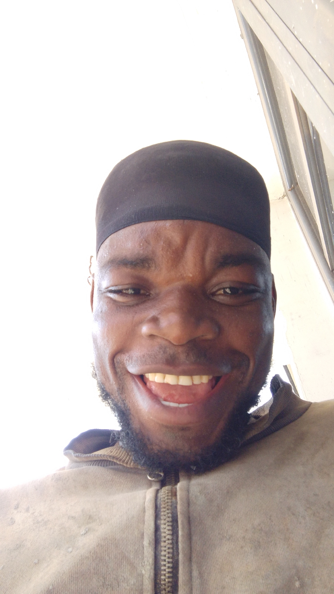 A close-up shot captures a smiling Black man wearing a black cap. He is wearing a zipped-up brown hooded sweatshirt. The background is predominantly white with a section of what appears to be a window frame. The location is Enugwu-Ukwu, Nigeria.