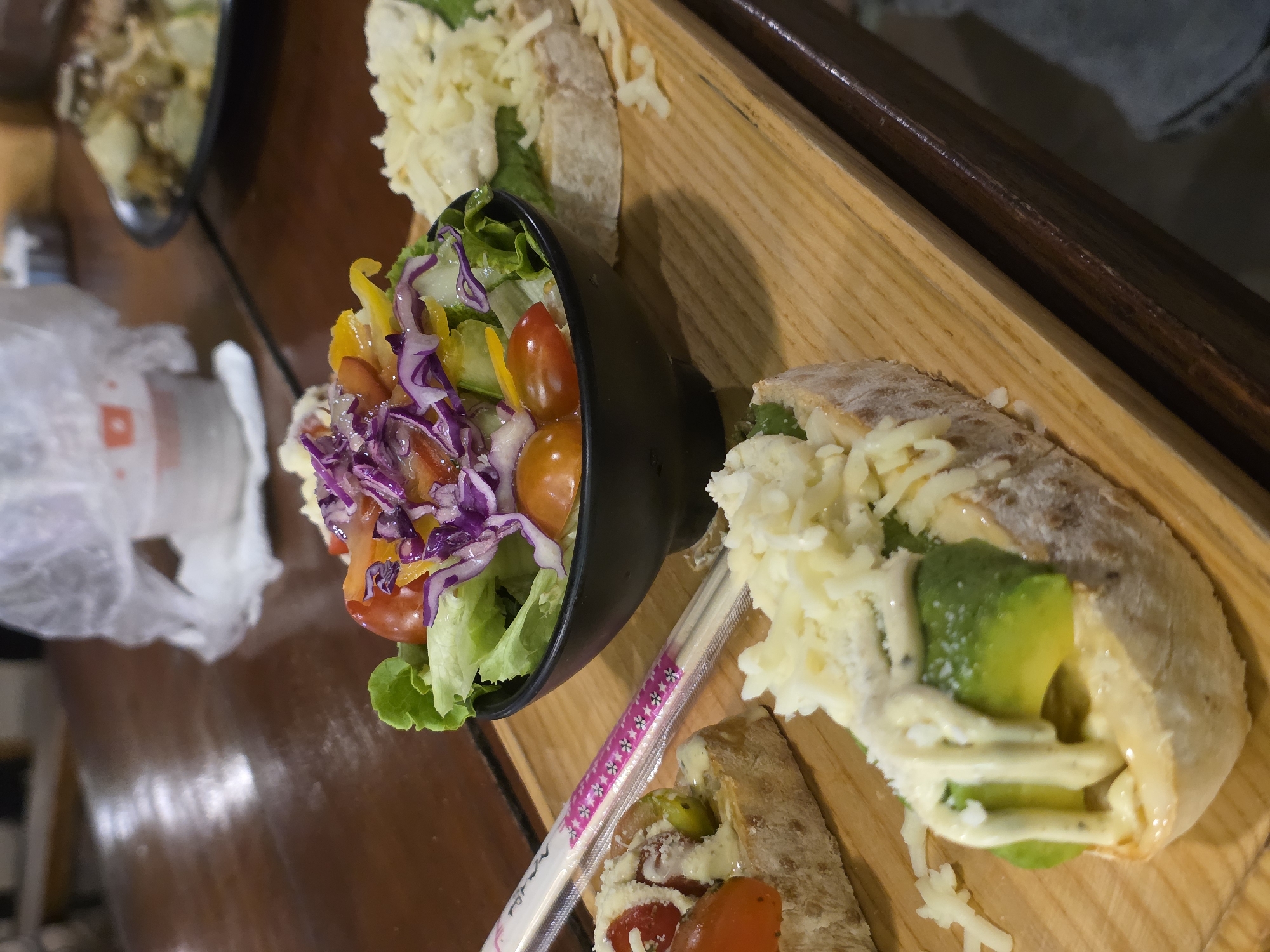 A close-up, eye-level shot captures a wooden platter holding various food items. In the center, a black bowl contains a vibrant salad with lettuce, cherry tomatoes, red cabbage, and yellow peppers, drizzled with dressing. To the left of the salad, a pair of chopsticks in a pink and white floral wrapper rests on the platter. In the foreground, a slice of bread topped with avocado, mayonnaise, and shredded cheese is visible, as is another similar slice in the background. The platter sits on a dark wooden table, and in the background, a blurry white cup and a plate of food can be seen. The setting is likely a restaurant in Phu Thuy Ward, Vietnam.