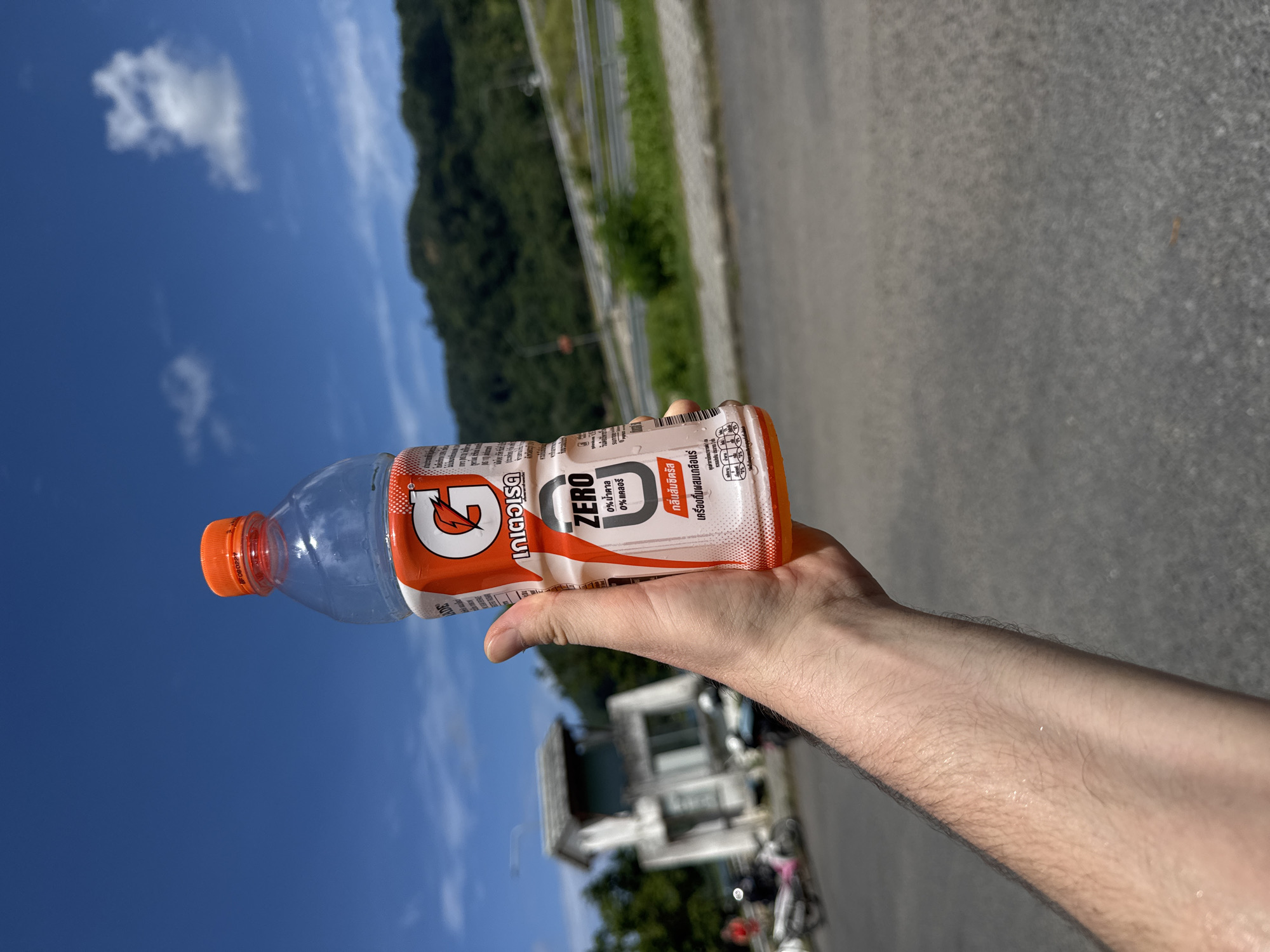 A Caucasian male is holding a bottle of Gatorade Zero. The bottle is orange and white with the Gatorade logo. The background is a blurry image of a road and a hillside covered in lush green trees. There is a small white building with a tan roof in the distance. The sky is a clear blue with a few white clouds. The man's arm is slightly sweaty, indicating a warm climate. The location is the city of Kathu, Thailand.