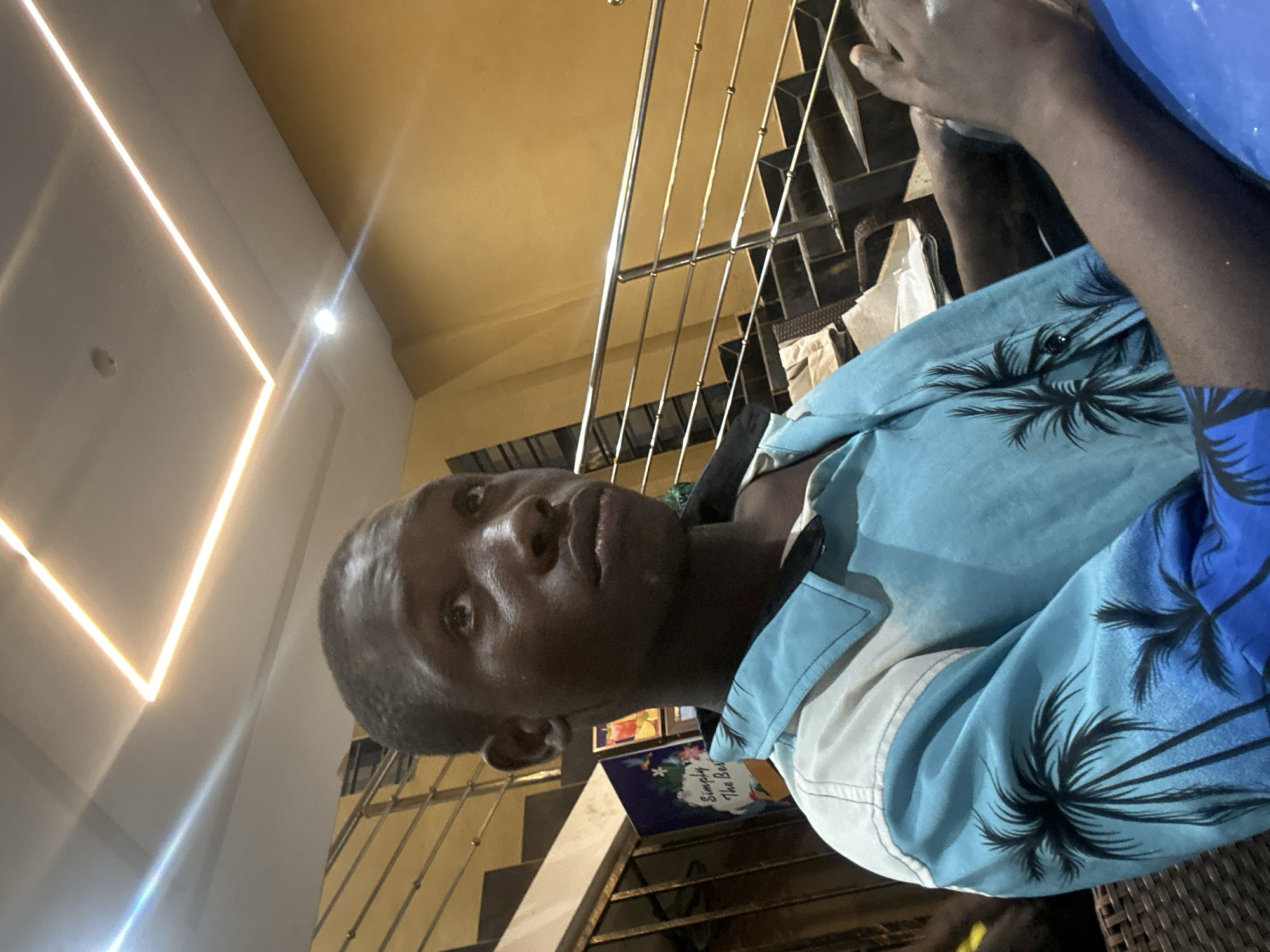 The image shows a young Black male sitting indoors, looking upwards and to the right. He is wearing a light blue short-sleeved collared shirt with a palm tree print. The background features a staircase with a silver railing and a yellow wall. The ceiling has a square recessed lighting fixture with illuminated strips. A sign with text and images is visible on the left side of the frame, near the stairs. The lighting suggests an indoor setting, possibly a restaurant or a public building. Ihiagwa, Nigeria, is a town in Imo State, known for its developing urban infrastructure.