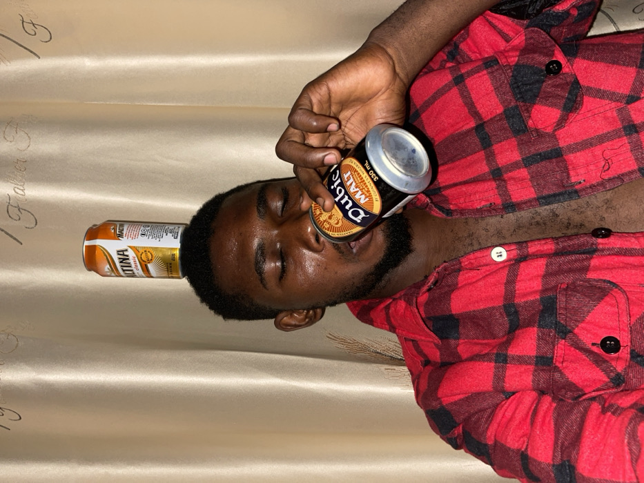 A man in a red and black plaid shirt reclines against a silky gold fabric. He has a beer can balanced on his forehead and another can pressed to his lips, seemingly drinking from it. The beer can on his forehead is orange, while the one he's drinking from is black with gold lettering. His eyes are closed, and he has a dark beard. The scene is casually staged and appears playful.