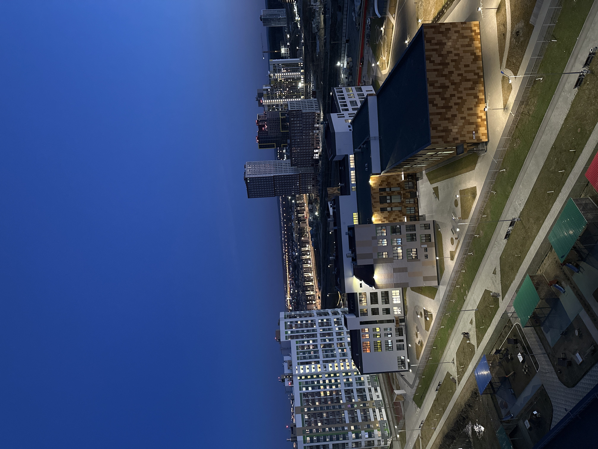The image presents a high-angle view of a modern urban development at dusk or night, characterized by a deep blue sky contrasting with abundant artificial lighting. The scene is dominated by multiple multi-story buildings, primarily residential, with numerous windows illuminated, suggesting occupancy.
In the foreground and mid-ground, well-designed contemporary buildings feature varied facades, including one with distinctive brown and light-toned square panels, and others with sleek white and grey sections. Winding pedestrian pathways, illuminated by numerous streetlights, connect these buildings and are flanked by manicured green spaces. In the lower right corner, several small covered structures with green and blue roofs suggest outdoor communal or play areas.
Further into the background, a dense cluster of high-rise buildings stretches towards the horizon, their lights creating a vibrant glow against the darkening sky. The overall impression is one of a newly developed or developing urban area.
There is no discernible human activity captured in the image. The sky is clear, indicating fair weather. No visible text or specific landmarks are identifiable within the frame. The artificial lighting from buildings and streetlights is a prominent feature, defining the urban environment during the blue hour or night.