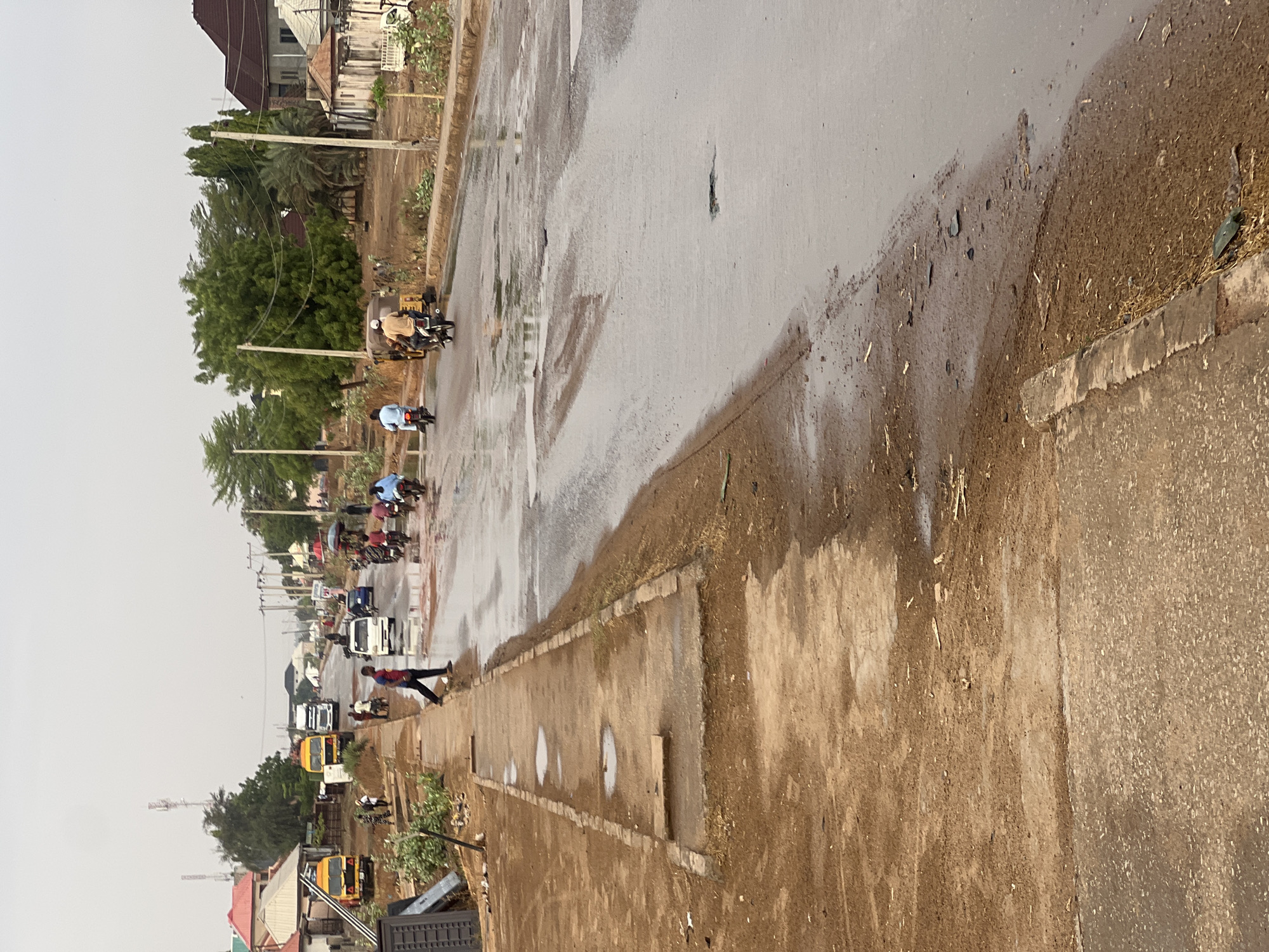 The image depicts a street scene in Minna, Nigeria, on a wet day. The road surface is slick with water. A line of vehicles, including motorcycles, cars, and buses, stretches into the distance. A pedestrian is walking along the sidewalk. Buildings and trees line the street. The sky is overcast.