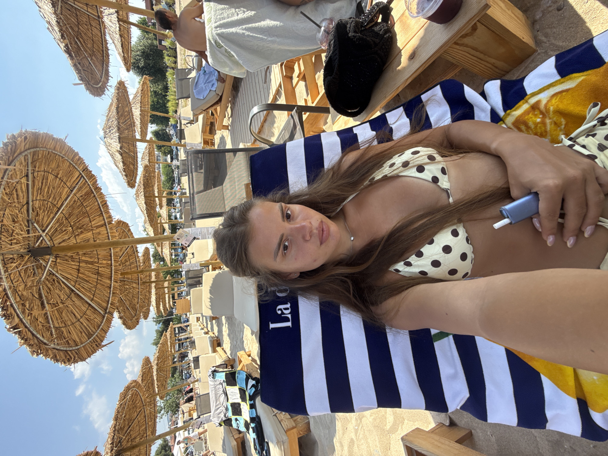 A young woman with long brown hair is lying on a striped beach chair at Ofrynio Beach, Greece. She is wearing a polka dot bikini and holding a small blue electronic device. The beach is lined with straw umbrellas and lounge chairs. In the background, other beachgoers are visible, as well as parked cars and trees. The sky is blue with scattered clouds.