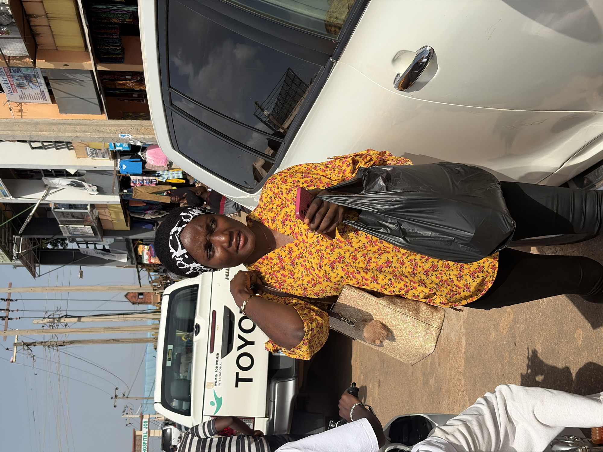 A Nigerian woman in a yellow floral print top and black pants stands on a dusty street in Jos, Nigeria, holding a pink cell phone and a black plastic bag. She wears a black and white patterned headscarf. To her left is the rear of a white Toyota pickup truck with "WOMEN FOR WOMEN INTERNATIONAL" and "TOYO" visible on its tailgate. To her right is the side of a white SUV. In the background, buildings with shops line the street, displaying clothing and other goods. The sky is clear and blue.