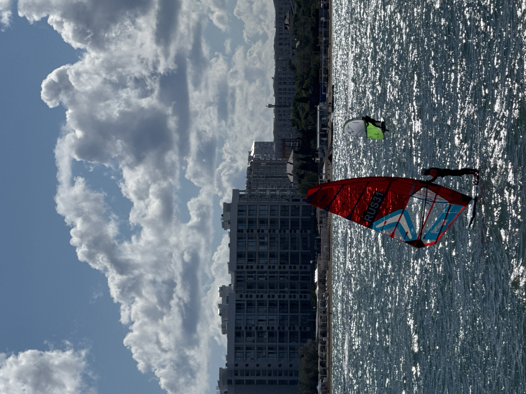 The image depicts a sunny day with dramatic clouds over a body of water, likely the Black Sea, with the city of Sevastopol, Ukraine, in the background. In the foreground, two individuals are windsurfing. One person is on a windsurfing board with a large, red sail that has blue and white patterns and the text "RUS31" visible. This individual is positioned in the lower right quadrant of the image. Further out in the water, a second windsurfer is visible with a smaller, green and white sail.
The middle ground shows a shoreline with a beach, dotted with white umbrellas and some vegetation. Behind the beach, a large, modern building complex with many windows dominates the cityscape. To the right of this complex, other buildings and structures are visible, including what appears to be a cell tower. The water is choppy and reflects the sunlight, creating a shimmering effect. The sky is a vibrant blue, filled with large, white, cumulus clouds, some of which are quite dense and cast shadows.