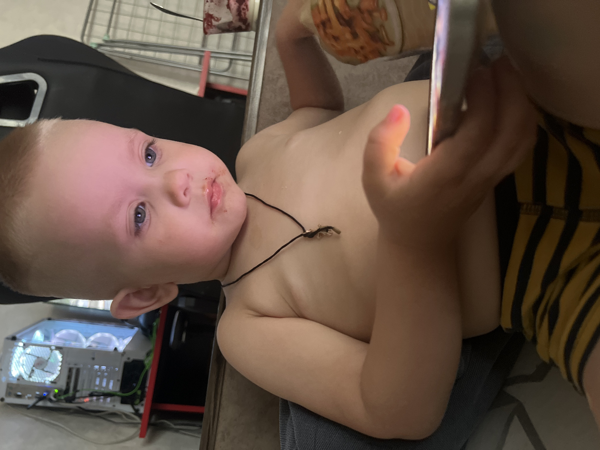 A young child, possibly a toddler, lies on their back, gazing at a phone held in their right hand. The child has fair skin, blonde hair, and blue eyes. There is food residue around their mouth. The child is wearing a necklace and a yellow and black striped garment on their lower body. The child appears to be on someone's lap, as blue denim material is partially visible beneath them.
In the background, there is a black object that appears to be a seat or piece of furniture, a metallic structure with a red bar, and a computer tower with illuminated fans. The location is Марковский, Russia, since that was the city that provided the context for analyzing the image.