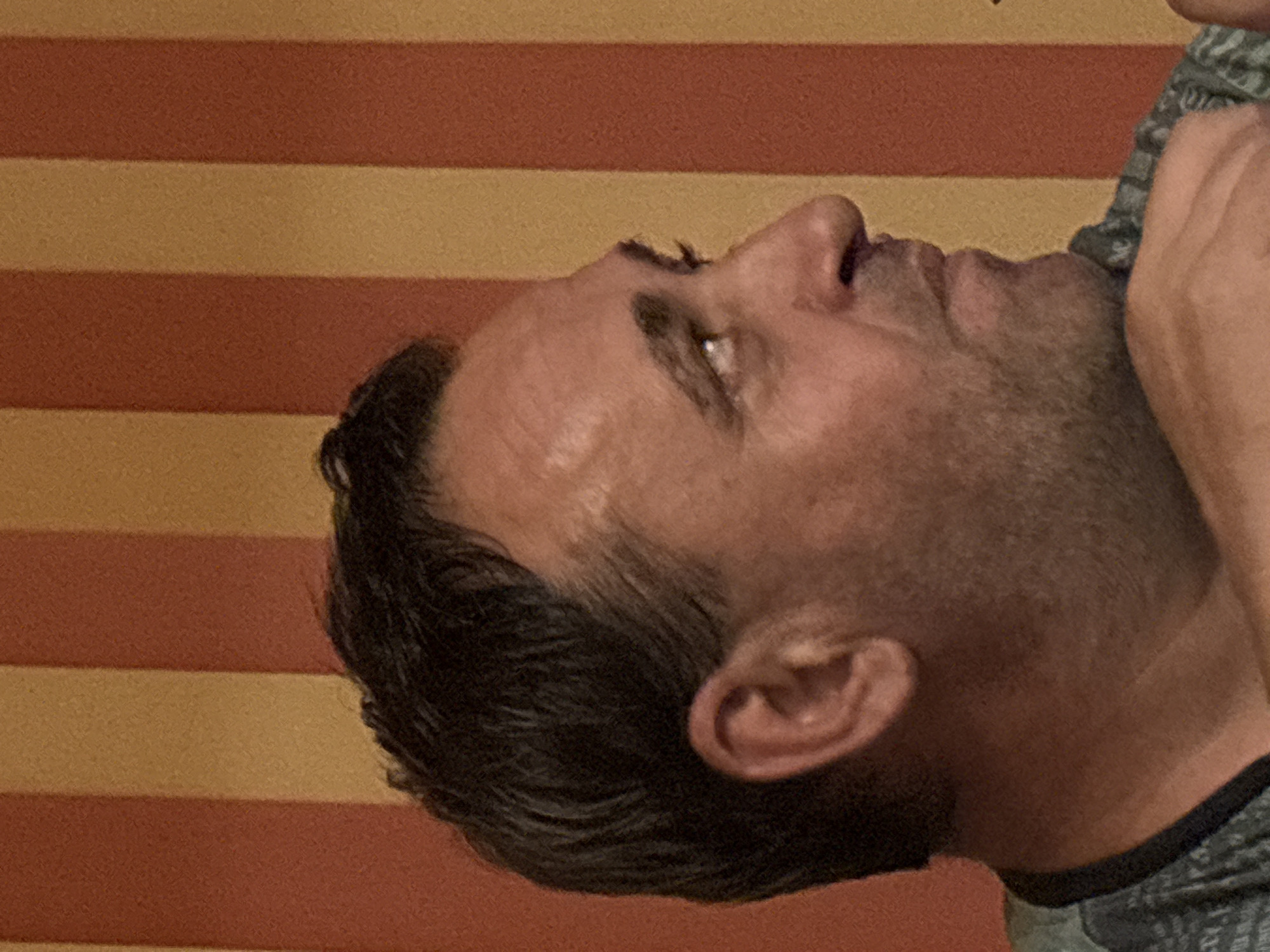 The image is a close-up, horizontally oriented, sideways shot of a person's head and neck against a background of horizontal stripes in shades of orange and yellow. The person appears to be male and is viewed from the side, with his head tilted upwards and to the right. His hair is dark and appears short, with some strands falling over his forehead. His skin is visible on his forehead, eye area, nose, cheek, and ear. The eye is open and looking upwards, with a glint of light visible. The nose is prominent, and the mouth is slightly open, revealing teeth. The ear is clearly visible and appears to be a light pink color. A portion of what appears to be an arm or shoulder is visible in the upper right corner of the image, with a patterned sleeve, possibly military-style camouflage in shades of green and grey. The lighting suggests an indoor setting with artificial light, possibly from the front, illuminating the face. There is no visible text in the image. The overall impression is of a candid shot, possibly taken during a casual moment, with no specific activity clearly depicted beyond the person being observed. The stripes in the background do not offer specific location cues beyond suggesting a patterned surface, possibly a wall covering or fabric. There are no indicators of time of day or weather conditions.
