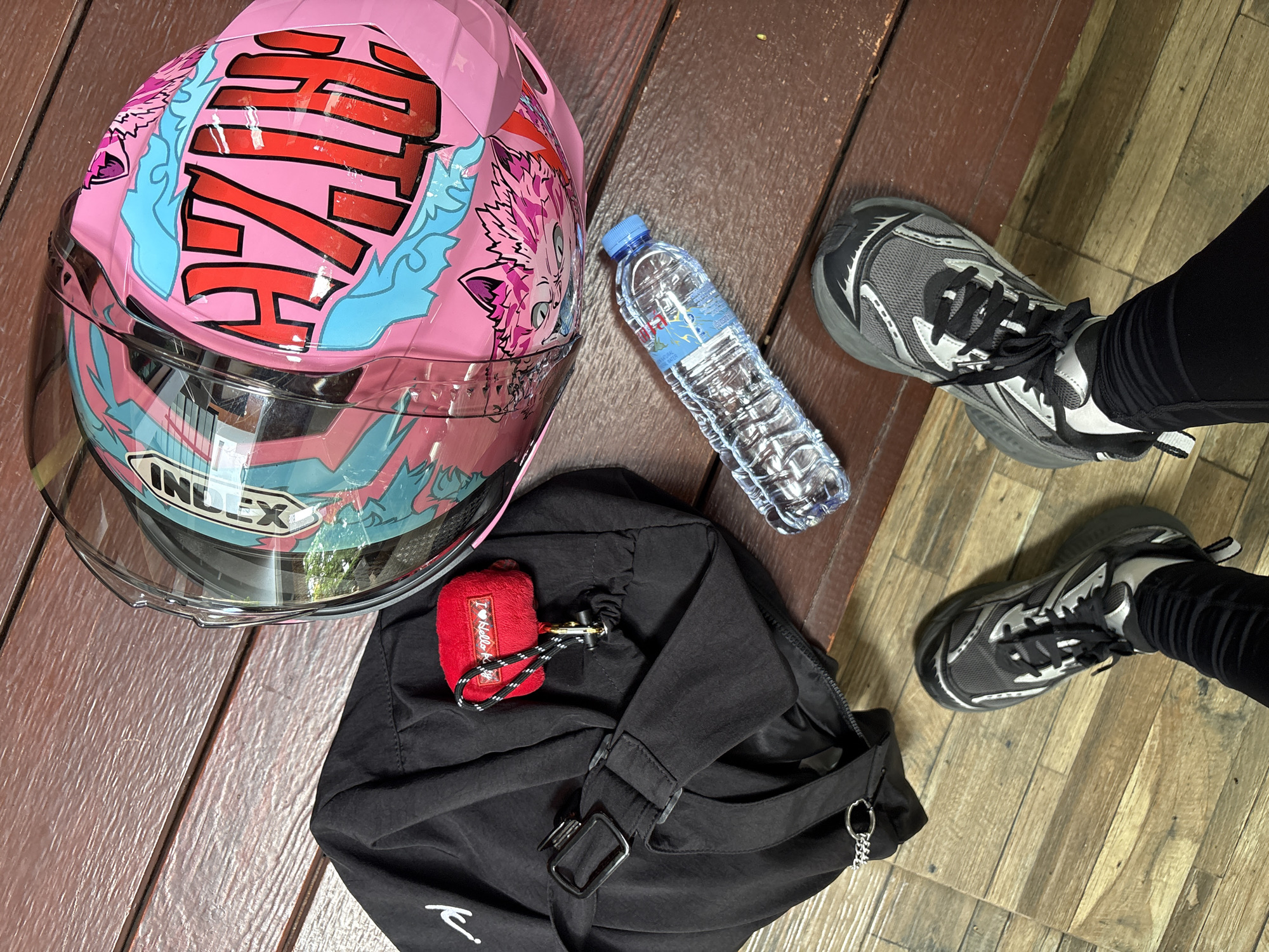 In the city of Ban Kan Chok 23, Thailand, a scene unfolds on a wooden deck. Two feet clad in black leggings and gray and black athletic shoes are visible, one positioned slightly ahead of the other. Nearby, a clear plastic bottle of water lies on its side. To the right, a black duffel bag is placed, adorned with a small red fuzzy pouch bearing the text "I <3 Hello K." A pink motorcycle helmet with a bold red and blue graphic design, featuring the word "CHZZH" in large red letters, rests in the foreground. The environment is an outdoor setting with a wooden floor.