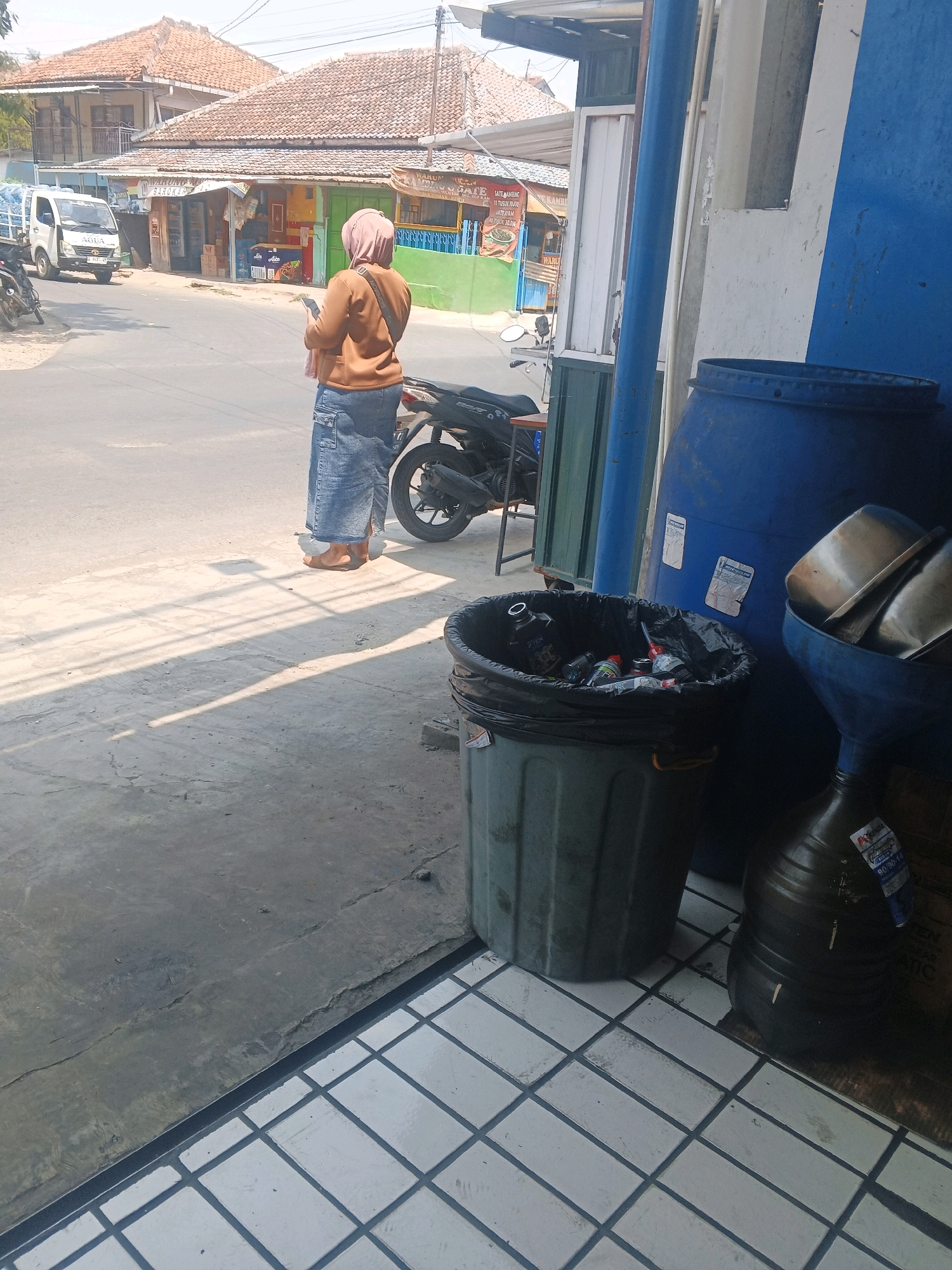 A woman stands on a street in Taman, Indonesia, facing a row of shops. She wears a brown hijab and a long denim skirt, and holds a smartphone in her hand. In the foreground, a dark-colored trash can is filled with various bottles and containers. Behind the trash can, a large blue barrel sits next to a funnel and another large container. The scene appears to be a commercial area with a paved road, shops with tiled roofs, and a white truck parked on the side of the road. The sun casts long shadows across the ground.
