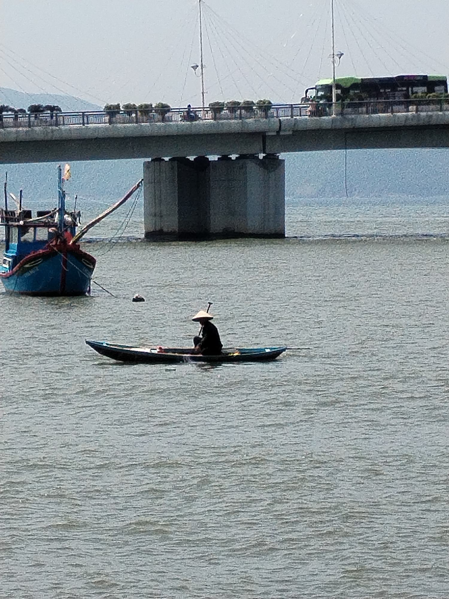 The image shows a wide, calm body of water with a small, dark blue boat in the foreground, occupied by a person wearing a conical hat and dark clothing, who appears to be fishing with a pole. To the left, a larger, blue fishing boat is anchored, displaying a yellow flag and some rigging. In the background, a substantial concrete bridge spans the water, with vehicles, including a green bus, visible on its deck. The bridge supports are massive concrete pillars. The water is a muted gray-blue with subtle ripples, and the sky is overcast. The scene appears to be set in a coastal or riverine area, likely in or near Bac Nha Trang Ward, Vietnam, given the presence of traditional fishing boats and a bridge indicative of urban infrastructure.