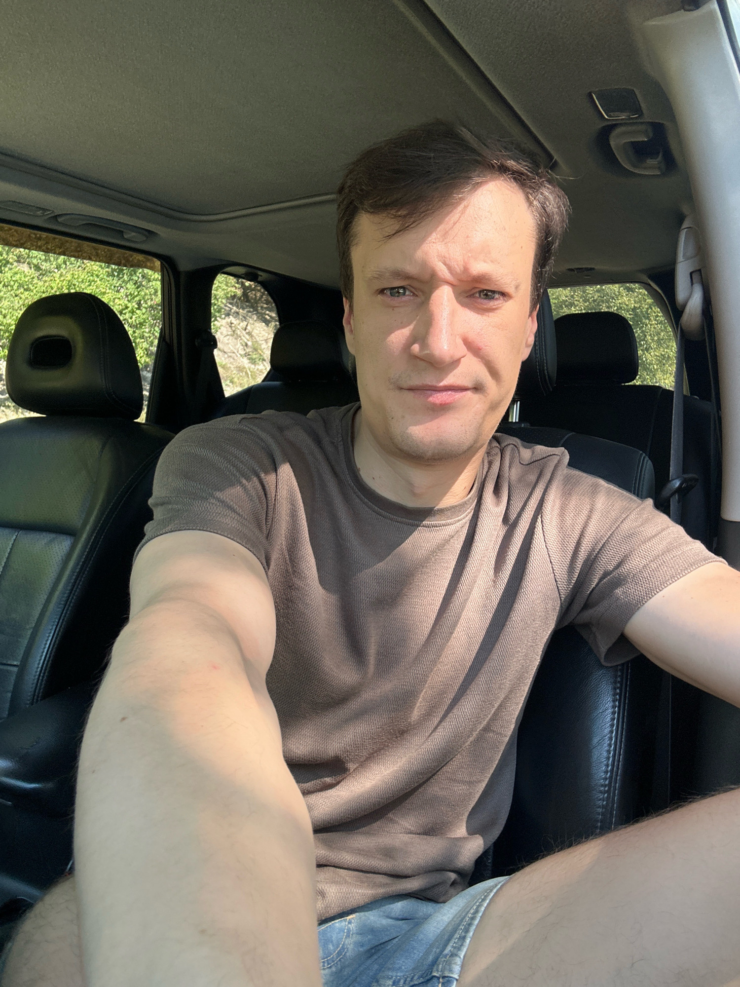 A man is sitting in the driver's seat of a car. He is wearing a brown t-shirt and denim shorts. The car's interior is visible, with black leather seats and a sunroof. Outside the car, greenery and trees can be seen, suggesting a rural or natural setting. The lighting indicates it is daytime. The image appears to be a selfie, taken by the man himself. The location is not specified in the image itself or accompanying information.