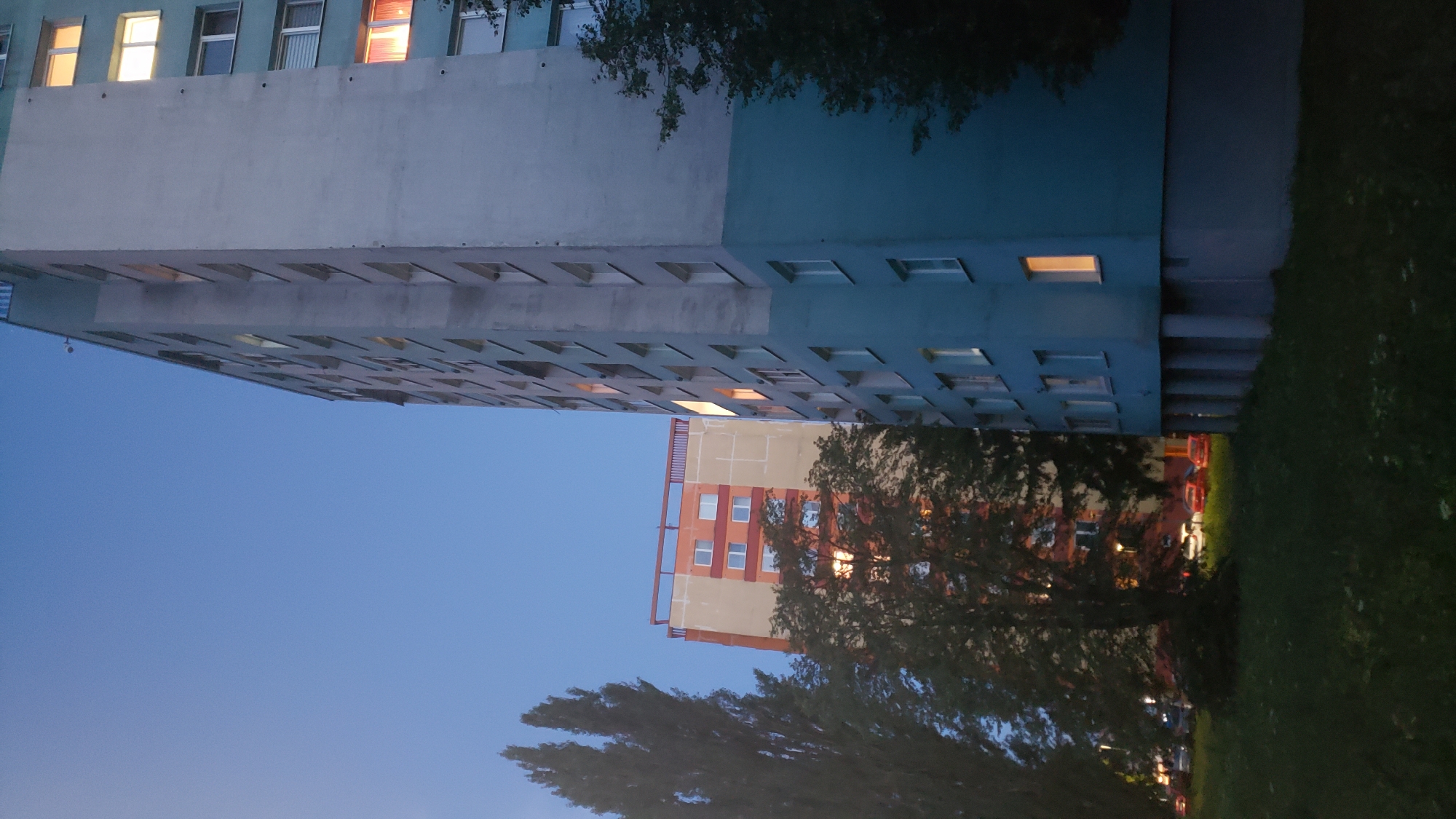 A low-angle, full shot captures an outdoor scene at dusk in Prešov, Slovakia, featuring two apartment buildings. The building on the right is a tall, pale grey structure with a blue base, featuring multiple floors with uniformly spaced windows, some of which are lit from within. To the left, a shorter, orange and yellow building rises behind a dense tree. This building also has multiple floors with windows, and its top section is adorned with a red railing. In the foreground, a grassy lawn stretches across the bottom of the frame, with several cars parked in the mid-ground between the buildings and across from a tree. Tall, slender trees are visible on the left side of the image, their dark silhouettes contrasting with the fading twilight sky.