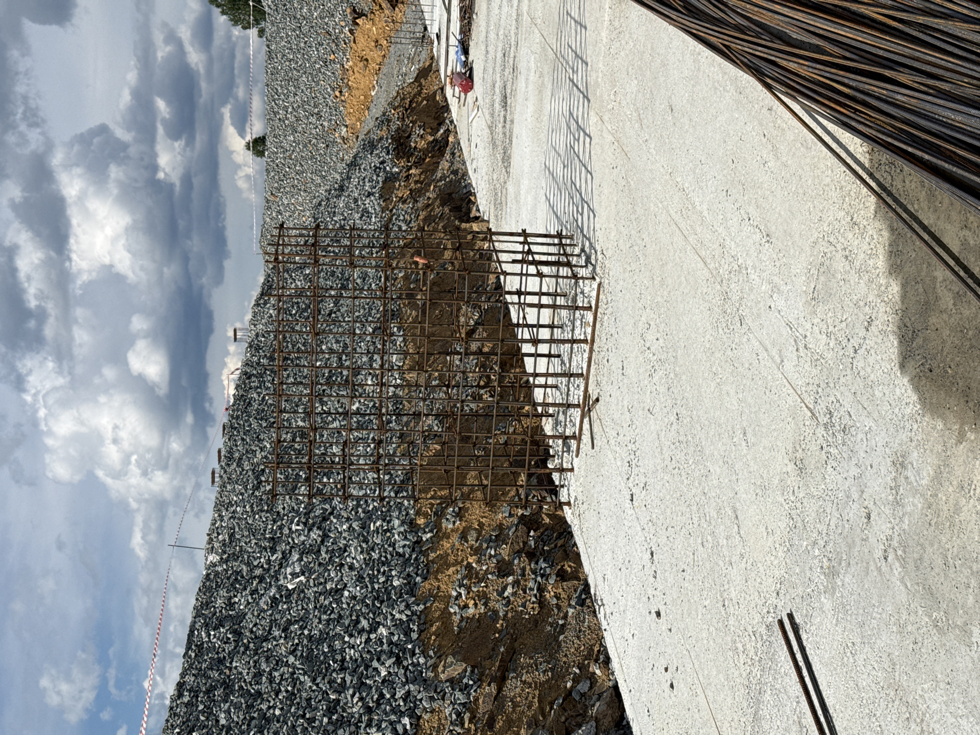 This is a full, eye-level, outdoor, daylight photograph of a construction site. In the foreground, a freshly poured concrete slab extends towards the background. A large, cube-shaped rebar cage stands in the middle ground, its metallic grid casting a shadow on the concrete. To the left of the rebar cage, a steep embankment composed of loose, grey gravel rises up, capped by a red and white striped caution tape. Further back, more gravel slopes down from the right side of the frame, with orange-brown soil visible beneath the gravel. The background features a partly cloudy sky with dramatic, dark clouds. On the concrete surface in the mid-right of the image, there are several construction materials, including a red circular object and some blue items, likely tools or safety equipment. Bundles of rebar lie on the concrete in the far right foreground. The scene depicts ongoing construction work, with the rebar cage indicating the preparation for a concrete pour or structural element. The location is identified as a construction site in the city of Leninsk, Russia.