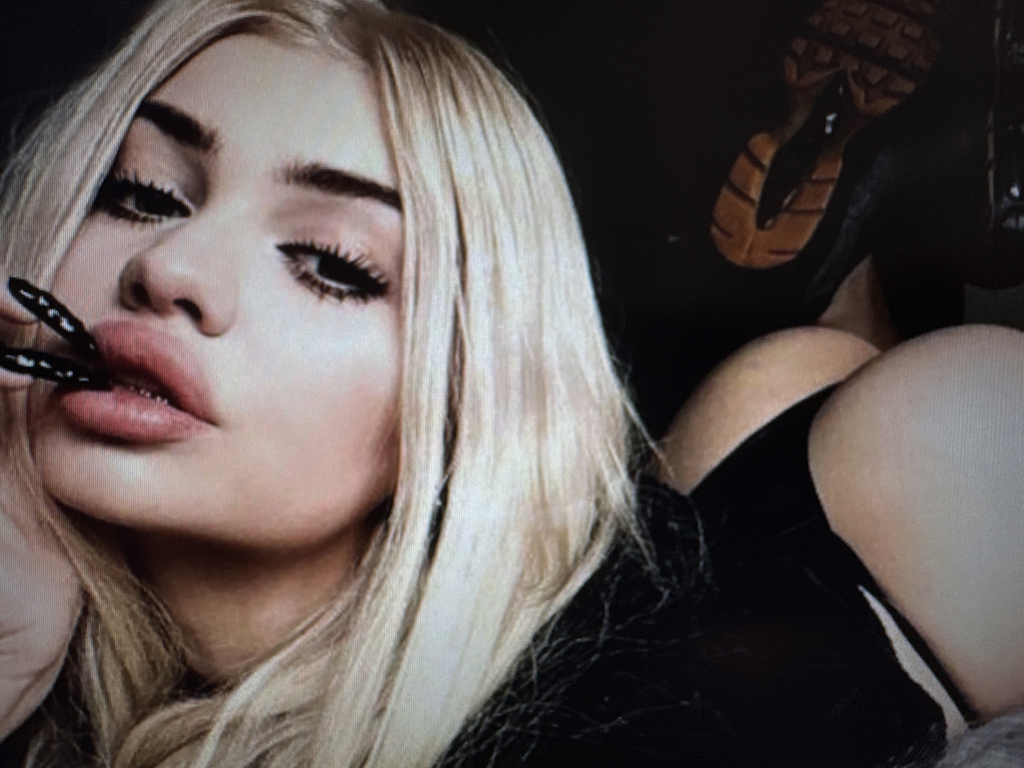This close-up image features a woman with long, straight blonde hair and prominent facial makeup, lying down and posing for the camera. Her face dominates the left side of the frame, showcasing dramatic winged eyeliner, thick eyelashes, and bold eyebrows. Her full, pouty lips are a focal point, highlighted with lip liner and a lighter shade. She holds a small, black, textured or spiked object near her lower lip.
Her body extends towards the right, with her right shoulder and a section of her buttocks, clad in black clothing or underwear, visible. In the upper right corner, the textured sole of a shoe, likely a high heel or platform, is partially visible against a dark background. The setting appears to be indoors and dimly lit, contributing to the intimate and somewhat sultry atmosphere. The image has a grainy quality, possibly indicating it's a photograph of a screen or taken in low light. The woman's expression is alluring and confident. No text is visible in the image. The provided location context is Varna, Bulgaria.