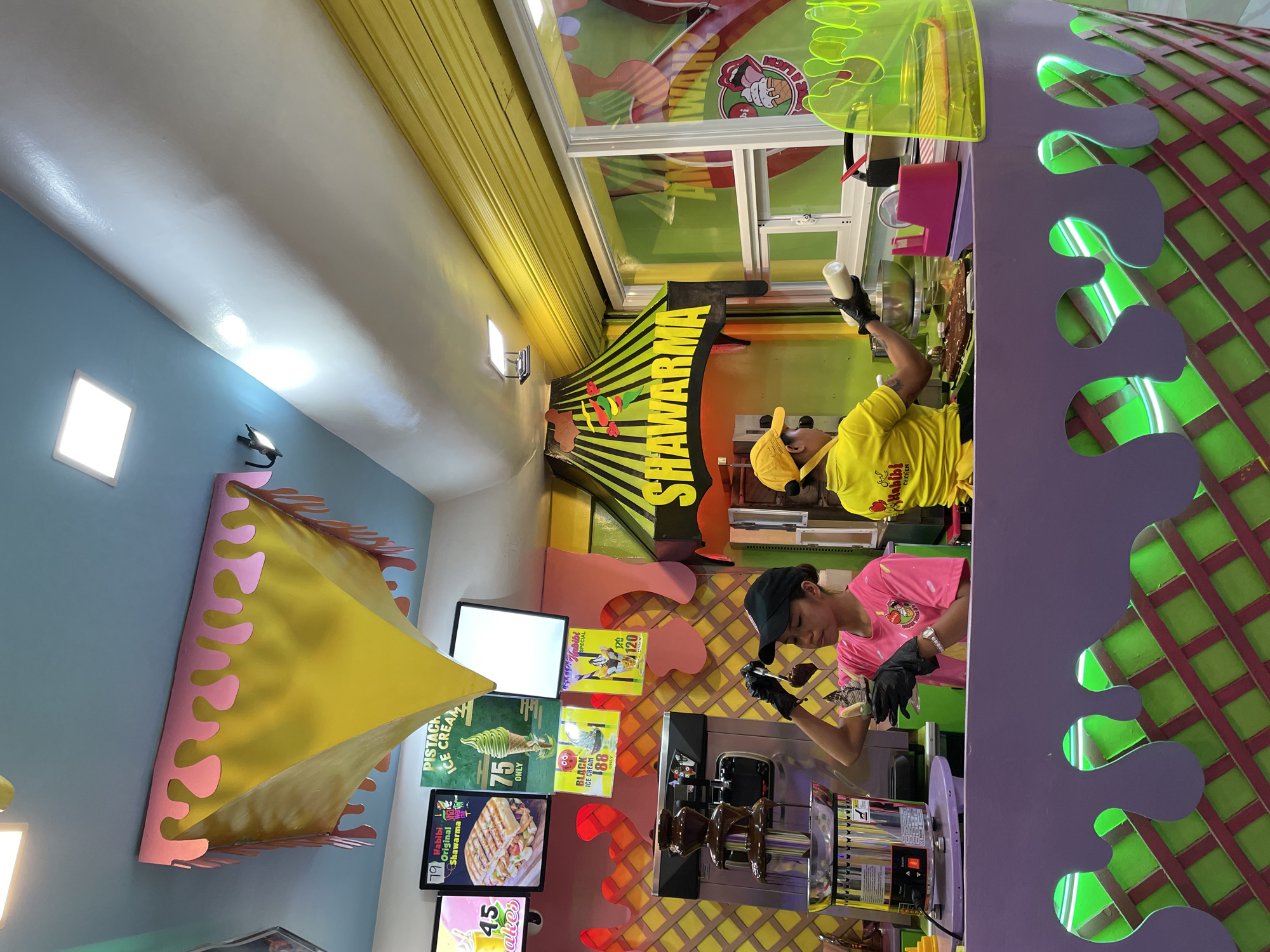 This image depicts a brightly colored food stall, likely located in Dumaguete, Philippines, specializing in shawarma and ice cream. The stall features a vibrant, playful decor with a yellow and pink ice cream cone-shaped canopy hanging from the ceiling, and a purple and green patterned counter. Two employees are visible: one in a pink shirt and black cap is dipping items into a chocolate fountain, and another in a yellow shirt and cap is preparing food behind the counter. Various signs advertise menu items, including "Habibi Original Shawarma" and "Pistachio Ice Cream." The overall atmosphere is lively and inviting, suggesting a popular spot for quick bites and treats.