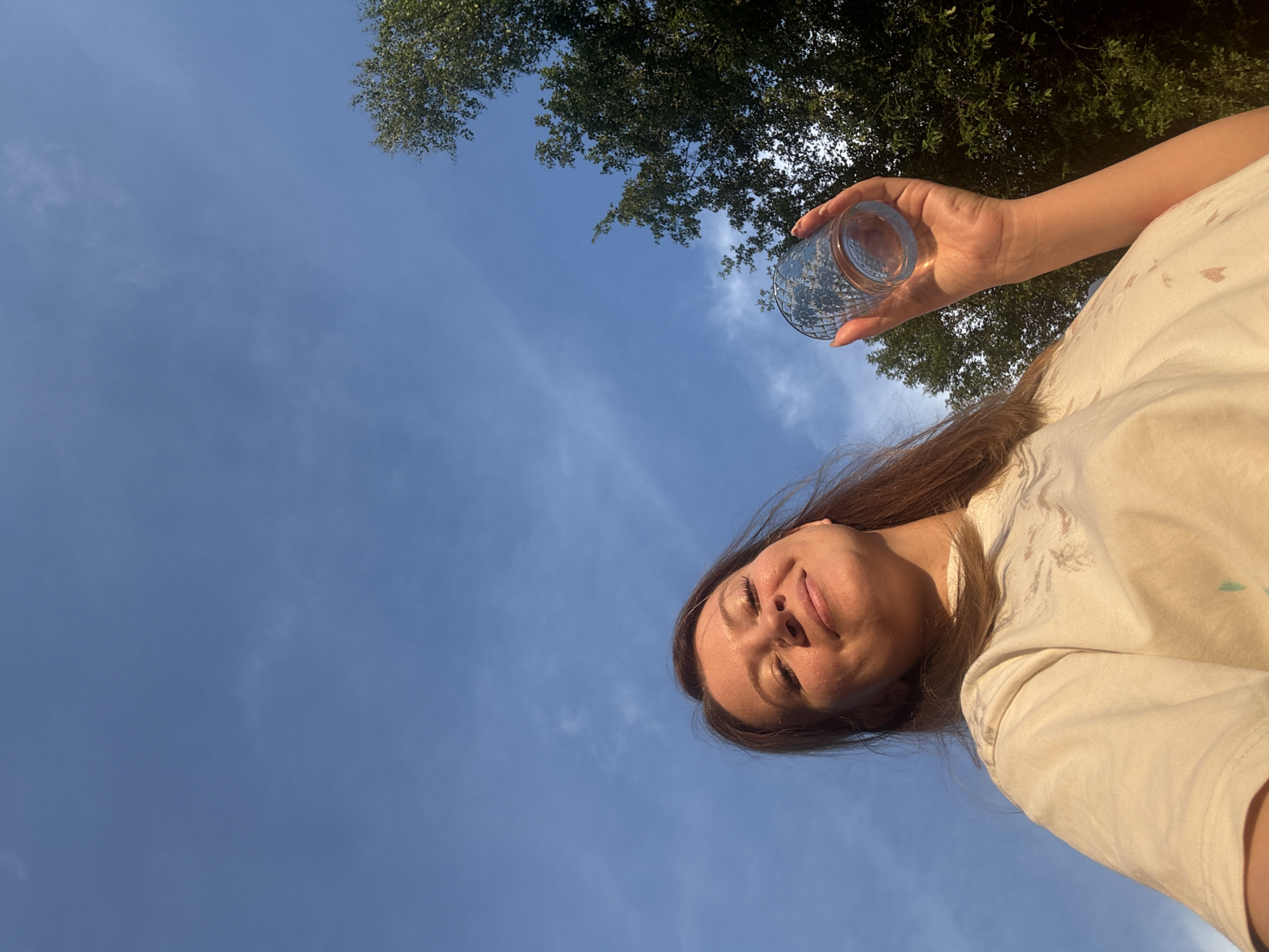 The image shows a woman holding a glass outdoors under a blue sky with scattered clouds. She is looking up and smiling, with trees visible in the background on the right side of the frame. The perspective is from below, looking up at the woman and the sky. The location is not specified in the image or metadata.