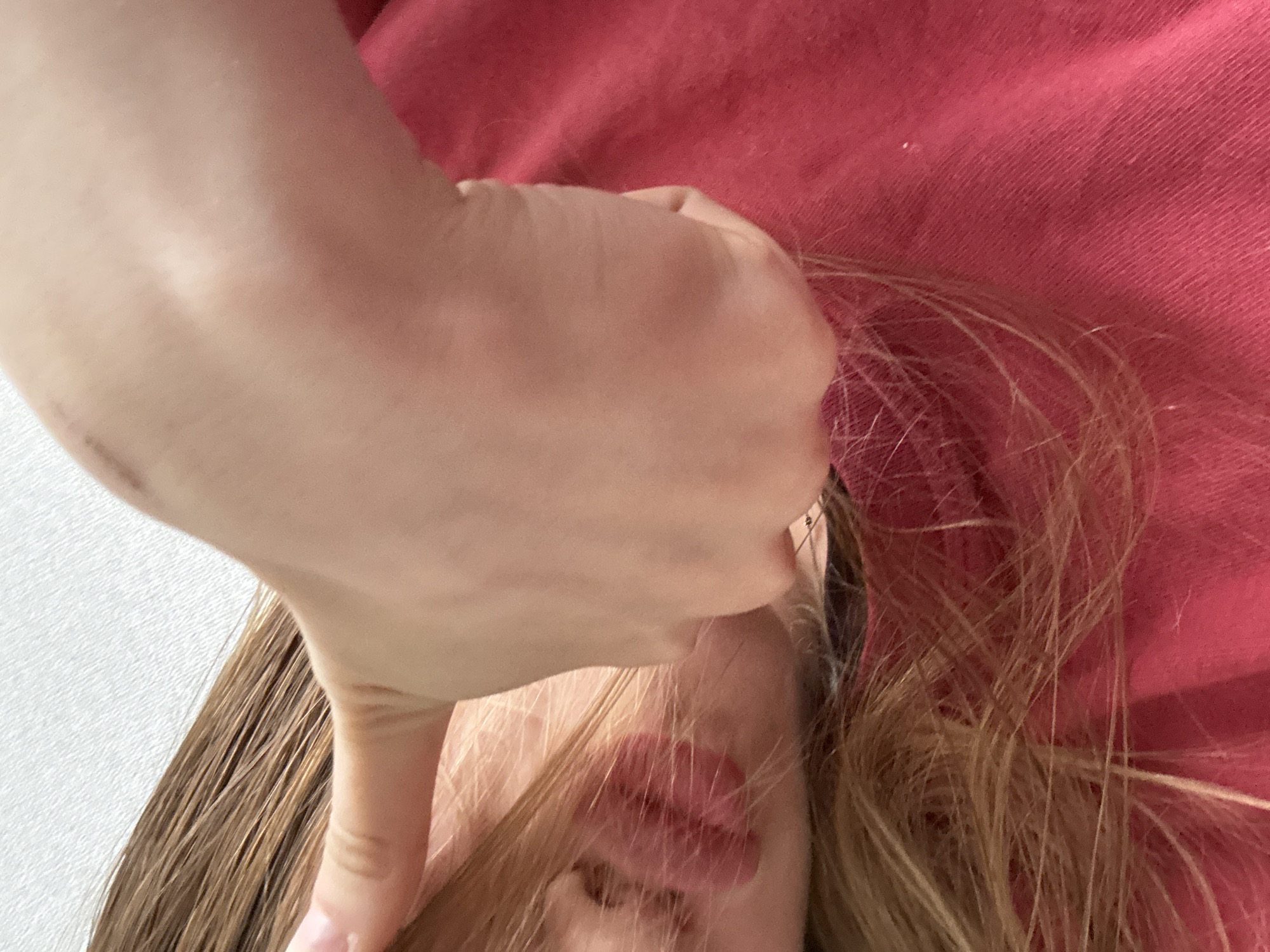 This media file is a close-up, overhead shot of a person's face and hand, with the person lying down. The person's face is partially obscured by their own hand, which is holding a strand of their hair. The hand appears to be the left hand of the subject, with light skin tone. The hair is light brown and straight, falling around the person's face and shoulders. The person is wearing a red garment, possibly a sweater or shirt, which takes up a significant portion of the background. The overall lighting suggests an indoor setting, possibly in daylight given the brightness. There are no visible signs of the time of day or weather conditions. The emotional state of the person is not clearly discernible due to the partial view of their face. There is no visible text in the image. The setting appears to be a home or a similarly private indoor environment. The image focuses on the interaction between the hand and the hair, creating a sense of introspection or a moment of personal care. The texture of the red garment is visible, with a subtle ribbed pattern. A small section of a textured white surface is visible on the left side of the image, which could be a wall or furniture.