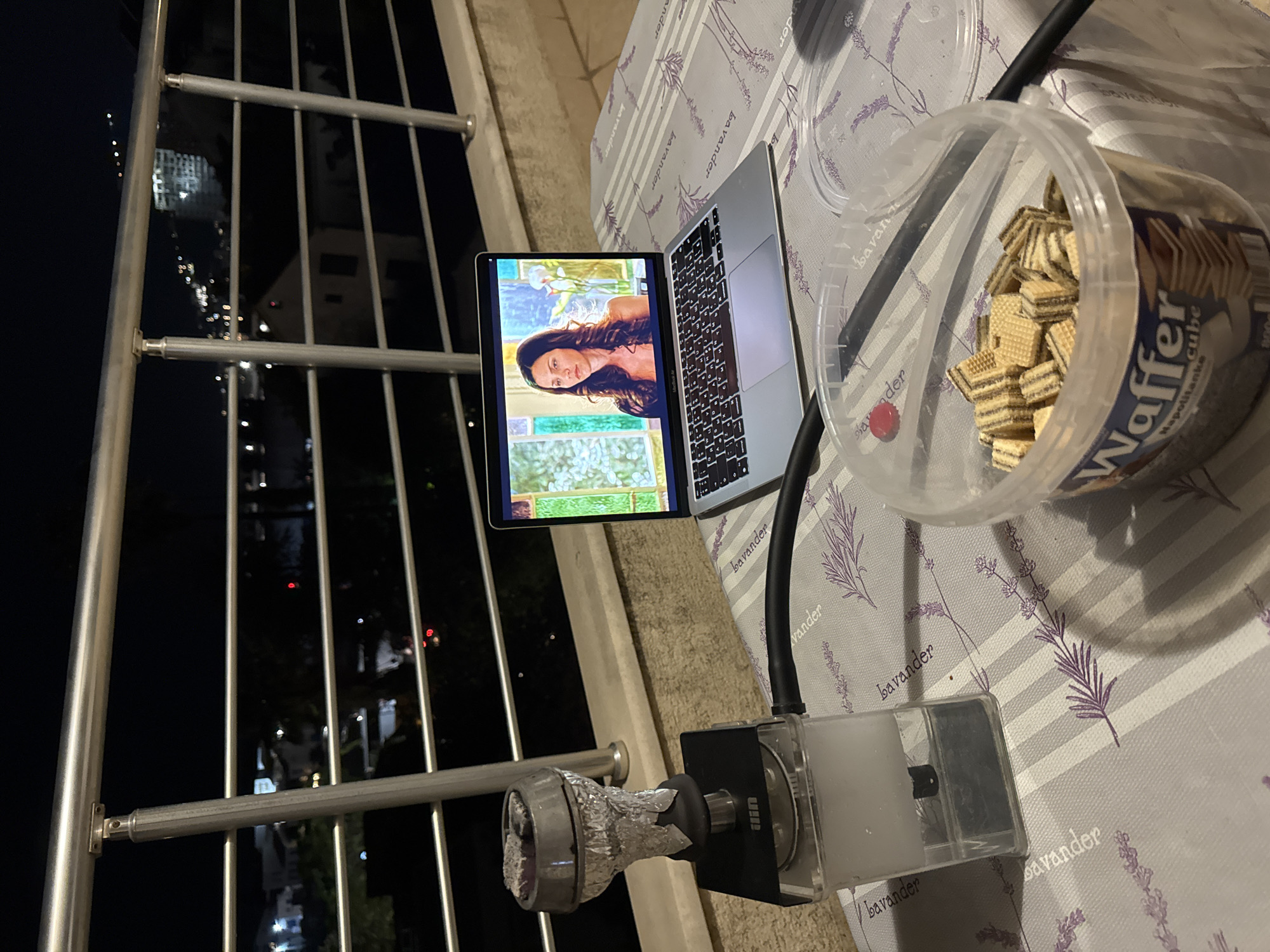 Here is a detailed description of the image:
**Scene Overview:** The image appears to have been taken on a balcony at night. The scene includes a laptop, a container of wafer cookies, and what seems to be a hookah. Through the balcony railing, city lights are visible in the distance.
**Objects:**
* A laptop is open, displaying a video or movie of a woman.
* A transparent plastic container filled with wafer cookies is on the table.
* A modern hookah is positioned nearby, consisting of a bowl wrapped in foil, a hose, and a water container.
**Location (Possible, based on visual cues):**
The city lights and buildings in the background suggest an urban environment.
Although not explicitly mentioned, the "Bavander" pattern on the tablecloth resembles the French word "Lavande" which means Lavender and also serves as a brand. Assuming this location is in Europe, it is possible that the scene is in Budva, Montenegro, or another coastal city with a similar urban setup.
**Environment:**
The scene is dimly lit. The balcony is furnished with a table covered with lavender-patterned fabric. The overall setting appears casual and relaxed, as if someone is enjoying an evening on their balcony.