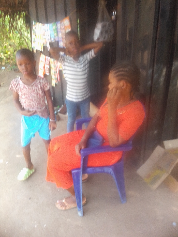 This image depicts three individuals—an adult woman and two children—in an outdoor or semi-outdoor setting in Eziudo, Nigeria, during the daytime.
To the left, a young child, likely female, stands wearing a patterned short-sleeved top and light blue shorts, with a single light green slipper visible on her left foot. Her gaze is directed towards the viewer. In the center background, another child, possibly male, stands with his arms raised behind his head, sporting a striped short-sleeved shirt and blue pants. Both children's faces are somewhat blurred due to the image quality.
To the right and in the foreground, an adult woman is seated on a bright purple plastic chair. She is dressed in an orange garment, possibly a dress or a top and skirt, and wears light brown sandals. Her hair is styled in prominent cornrows, and her left hand is positioned near her face, suggesting a moment of rest, thought, or observation.
Behind the children, against a dark, possibly wooden wall, a string or line holds numerous small, colorful rectangular packets, which appear to be items for sale, such as snacks or small goods. A clear plastic bag containing darker items hangs from above in the background. The ground is a light, unpaved surface, and some green vegetation is visible in the far-left background, indicating a natural environment beyond the immediate structure. The scene portrays a casual, everyday gathering or activity. No legible text is visible in the image.