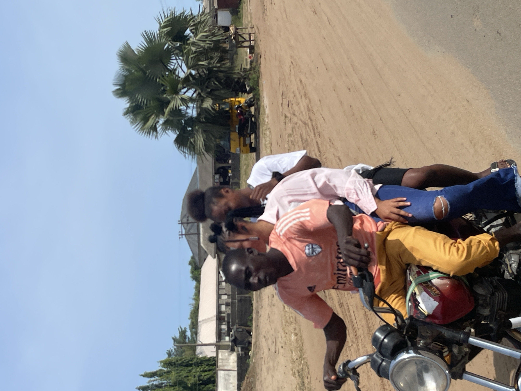 A motorcycle is moving down a dusty road in Ipota, Nigeria. The motorcycle is ridden by a young man wearing a peach-colored shirt and yellow pants. There are two female passengers behind him. One female passenger is wearing a white shirt, the other a pink shirt and jeans with a hole in the knee. A red helmet is on the motorcycle. In the background are buildings and a palm tree.