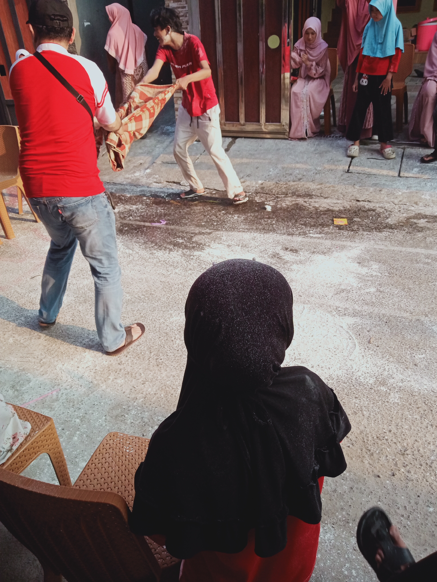 In East Jakarta, Indonesia, a scene unfolds on a concrete ground surface where several individuals are present. In the foreground, a person wearing a black hijab and a red garment is seated on a brown plastic chair, their back to the camera. To their left, another brown plastic chair with a floral patterned cloth is visible. In the middle ground, two men are engaged in an activity involving a patterned blanket. One man, wearing a red shirt and jeans, holds one end of the blanket, while another man in a red shirt and white pants pulls on the other end. Behind them, several women in hijabs of pink and blue are standing or sitting on chairs. The ground is scattered with white powder and some wet patches. The environment appears to be an outdoor space, possibly a residential yard or a street.