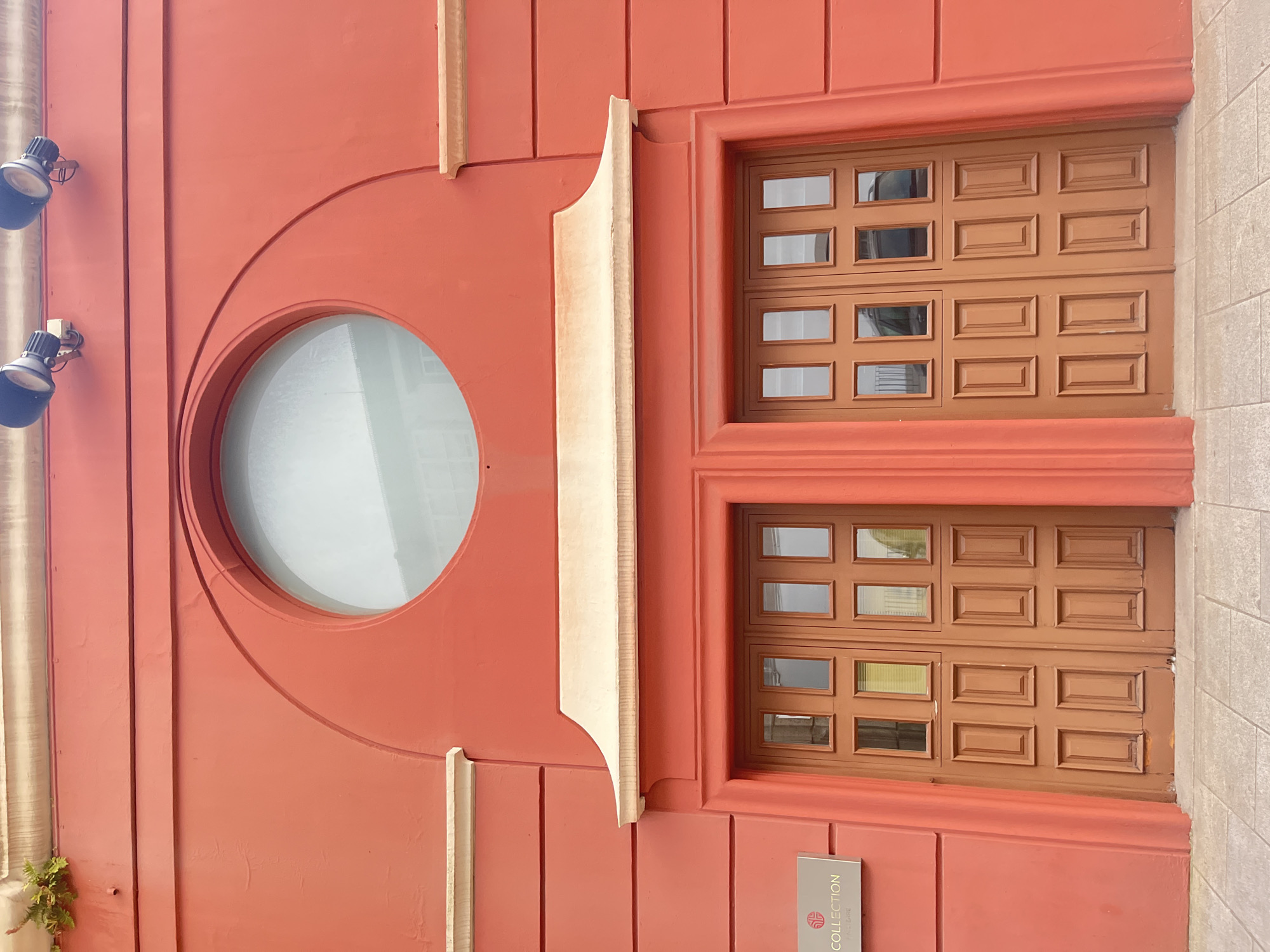 The image shows the exterior of a building in A Coruña, Spain. The building has a bright orange facade and features a large circular window with a light-colored frame. Below the window, there is a decorative architectural element resembling a cornice, also in a light color. Centered below this element are two large wooden double doors, each divided into multiple rectangular panels. To the left of the doors, mounted on the orange wall, is a silver plaque with a logo and the text "COLLECTION". The building's facade is textured and has horizontal decorative lines. Two spotlights are mounted on the upper part of the facade, pointing downwards. The ground in front of the building is paved with light-colored square tiles. A small patch of green vegetation is visible on the top left corner of the facade. The scene appears to be an entrance to a commercial establishment.