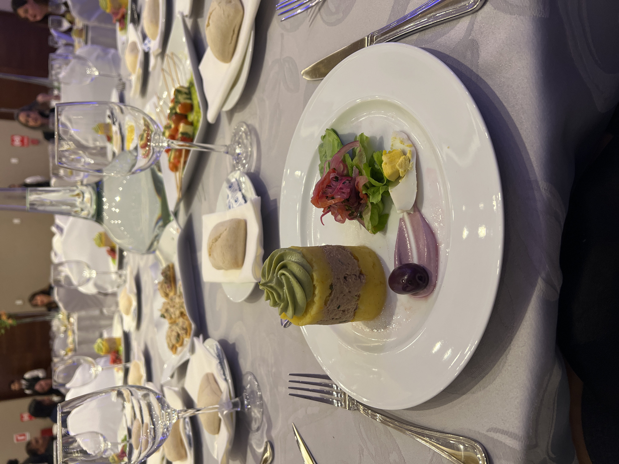 This image depicts a formal dining setting, likely a restaurant or banquet hall, at a table covered with a grey tablecloth with a subtle pattern. In the foreground, a white plate holds a meticulously arranged dish consisting of a cylindrical tower of food topped with green puree, a layer of meat filling, and a base of yellow puree. Beside the tower is a small salad with red onions and leafy greens, a halved hard-boiled egg, and a dark olive on a swirl of purple sauce. A fork and a knife are placed to the left and right of the plate, respectively.
In the background, several other tables are visible, set with white plates and glassware, indicating a meal in progress or about to be served. People are seated at some of the tables, though their faces are not clearly discernible. Wine glasses, some empty and some with reflections, are prominent on the tables. A large glass carafe is also visible. The overall ambiance suggests a special occasion or a fine dining experience.
Without further information or contextual clues within the image itself, pinpointing the exact location within the city of Arica, Chile, is not possible. The scene is consistent with upscale dining establishments found in any major city.