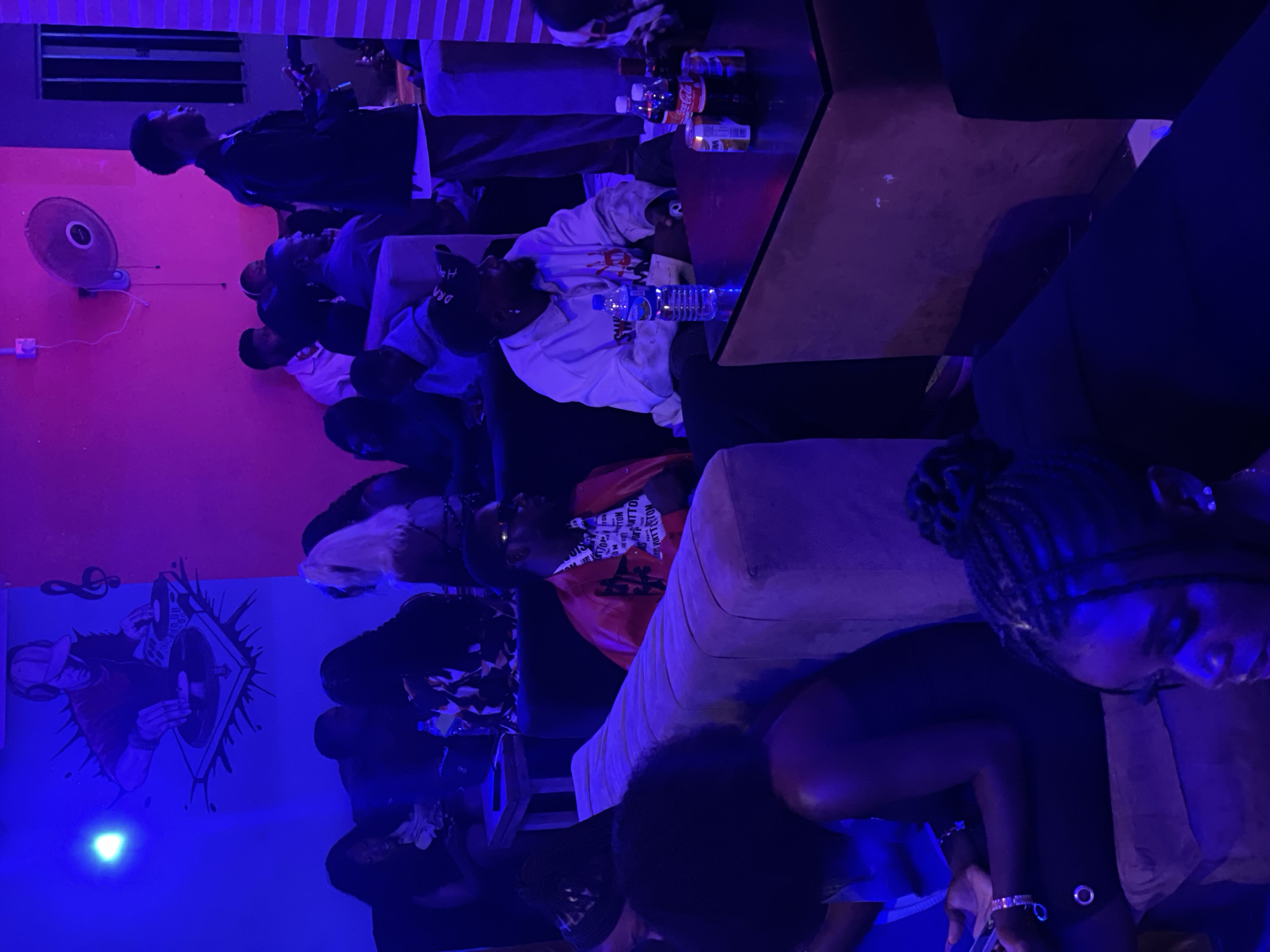 The image depicts a dimly lit, indoor space, likely a lounge or club, with vibrant blue and pink lighting. The walls feature a mural of a DJ and musical notes on the left, and a fan on the upper right. Several individuals are seated on couches and chairs, engaged in conversation or observing their surroundings.
In the foreground, a woman with braided hair is partially visible on the right, looking down. To her left, a person in a bright red jacket with intricate white patterns on the inner lining is seated. In the mid-ground, a man in a white hooded sweatshirt with handprints and text on it is seated, with a bottle of water and cans on the table in front of him. Other individuals are scattered throughout the scene, some partially obscured by the low light and framing of the shot. The overall atmosphere suggests a social gathering. The location is identified as Durumi, Nigeria.