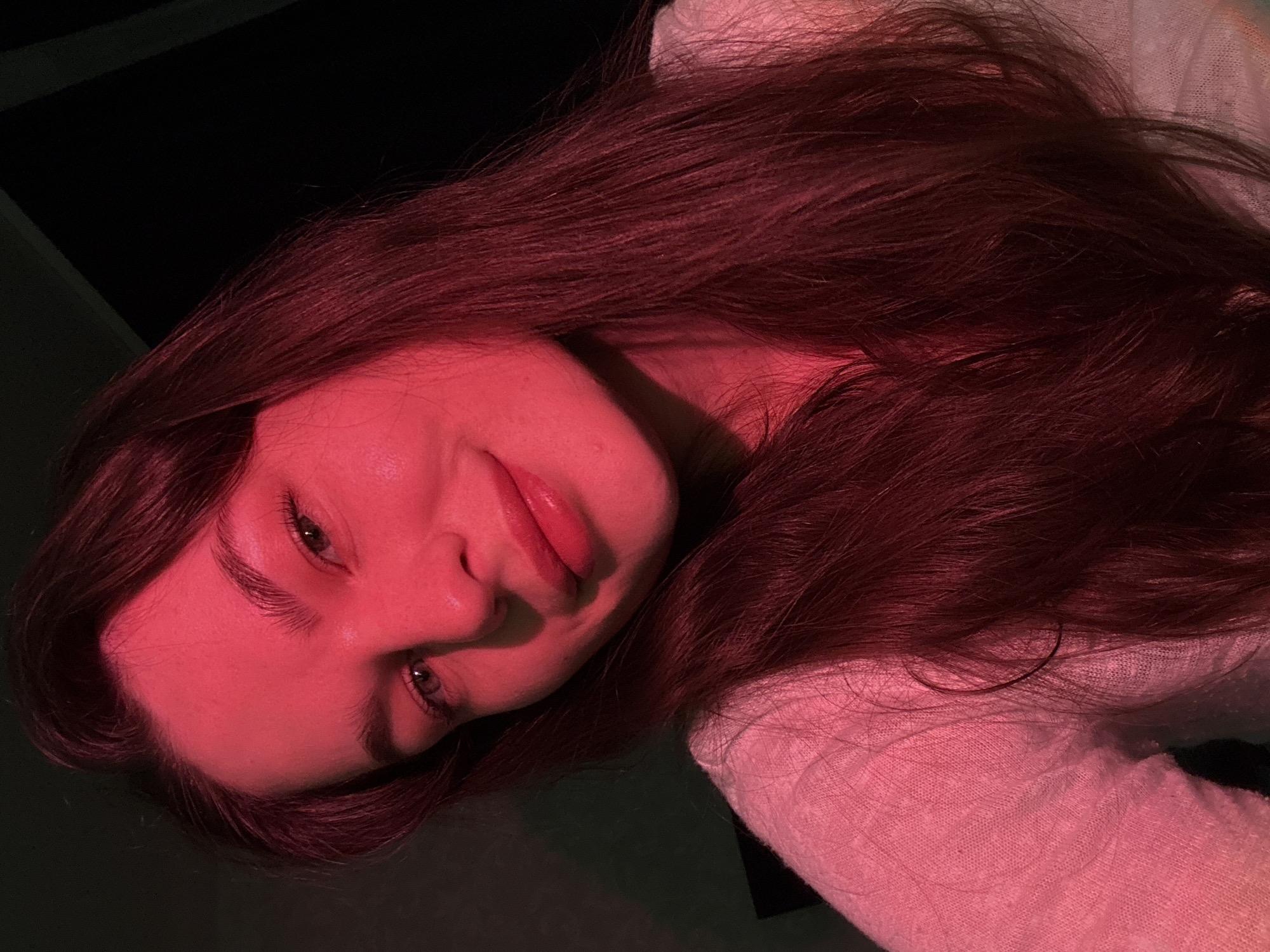 A young woman with long, dark hair is looking at the camera. She has on a light-colored sweater. The lighting is predominantly red. The background is dark. The image appears to be a selfie. The location is not specified in the image and cannot be determined from the visual information.