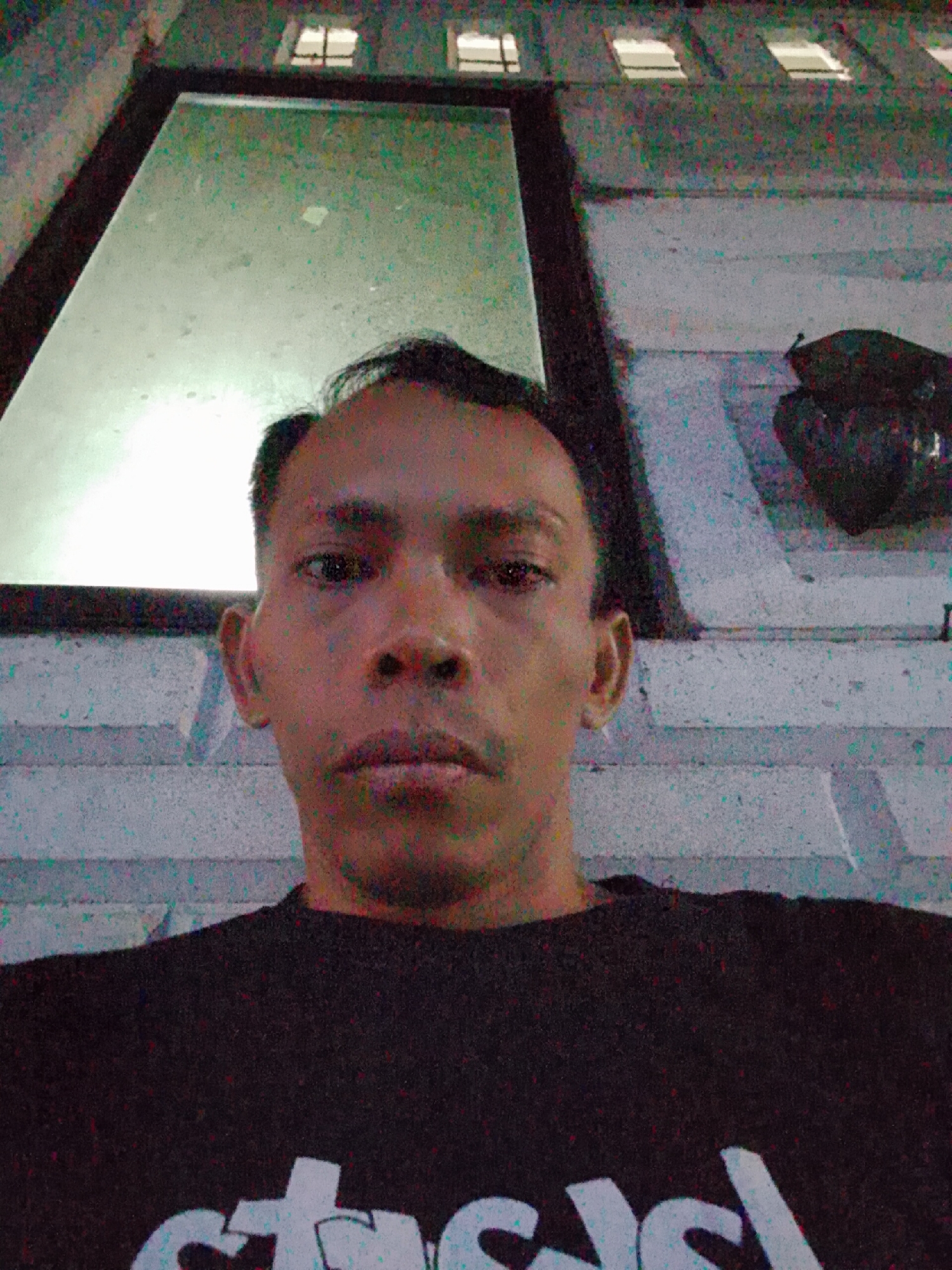 A low-angle selfie of an adult male, likely Indonesian, positioned in front of a building in Bukit Ketok, Indonesia. The male has short, dark hair, brown eyes, and is wearing a dark-colored t-shirt with white lettering on the lower part of the frame. Behind him, the building features a large window and possibly a shutter or corrugated metal section along the top with rows of small rectangular window openings. There is an object, potentially a bag or some folded material, hanging on the wall to the right of the window. The lighting is somewhat dim and artificial.