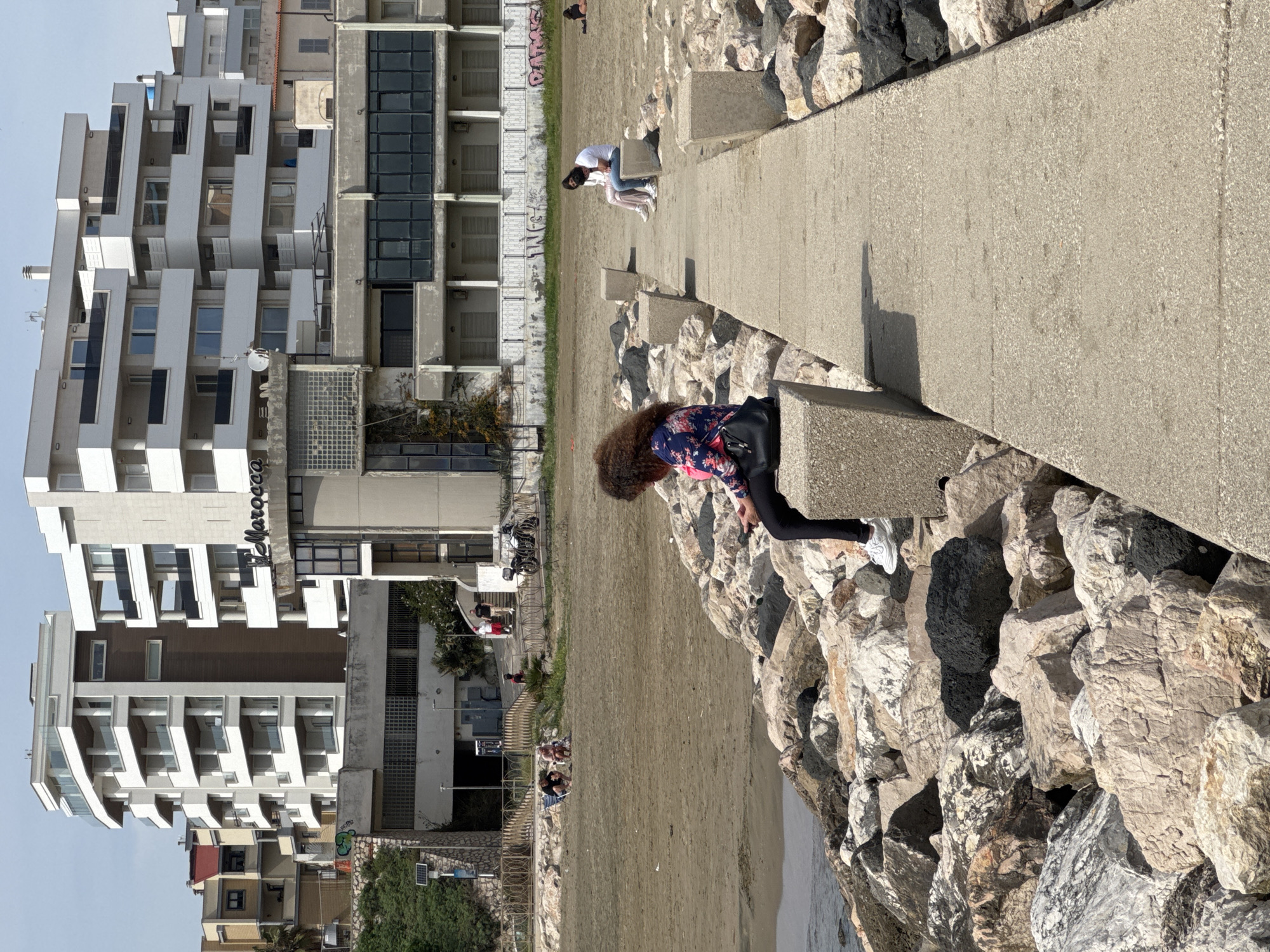 The image captures a scene at Nettuno, Italy, showing a beachfront setting with concrete structures and buildings in the background. The foreground features a woman with curly hair leaning over the edge of a concrete pathway or pier, looking down towards rocks and the water. She's wearing a floral-patterned shirt and black leggings. Another person is sitting further along the path. The background includes modern buildings with white balconies and a mix of architectural styles. The beach is sandy with people relaxing and walking in the distance. The sky is light blue with some clouds.