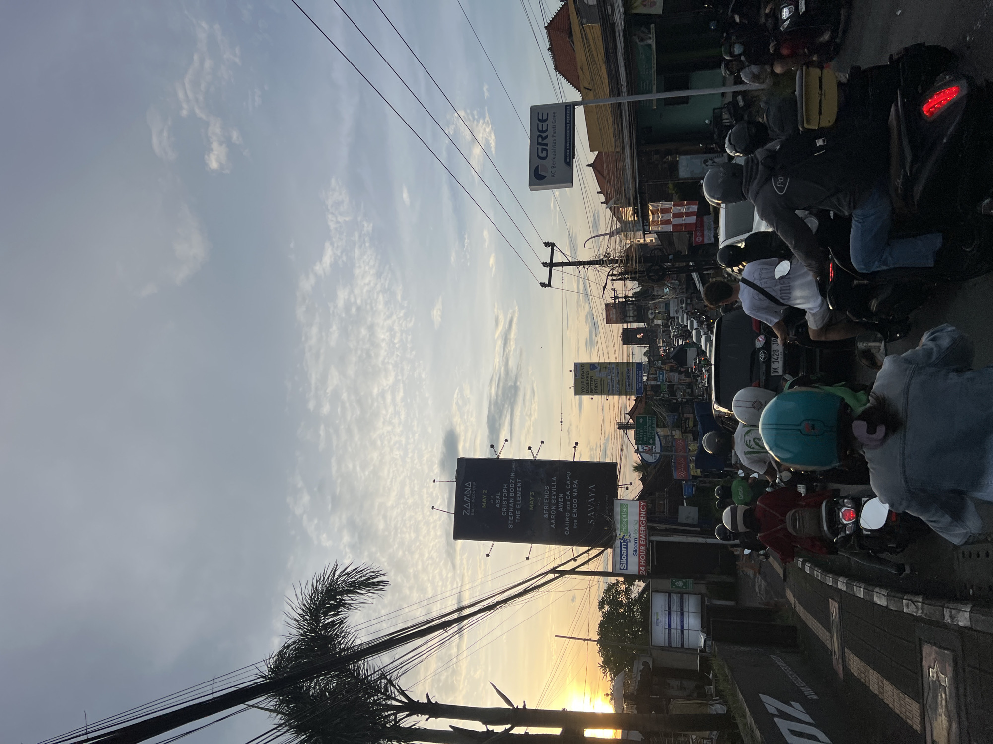 The image captures a busy street scene in North Kuta, Indonesia, at sunset. The sky displays a soft blue with scattered white clouds, transitioning to a warm golden glow from the low sun on the right horizon. Utility poles and a dense web of overhead power lines are prominent across the upper frame.
The street is active with significant motorbike traffic, showing numerous riders, many wearing helmets, either stationary or moving slowly in congestion. Buildings line both sides of the road, featuring diverse architecture and commercial establishments. A large palm tree is silhouetted against the bright sky on the left.
Visible text on various signs and billboards provides location and commercial details. A large black billboard advertises "ZAMNA INDONESIA" with event details for "MAY 2" and "MAY 3" featuring artists like "ASAL," "CRISTOPH," "STEPHAN BODZIN (LIVE)," "THE ELEMENT," "AARON SEVILLA," "AWEN," "CAIIRO," and "ENOO NAPA" at "SAVAYA." Another sign advertises "GREE AC Berkualitas Pasti Gree MITRA ENGINEERING PERKASA." A green sign indicates "Siloam Hospitals Siloam Medika 24 HOUR EMERGENCY." A partial sign reads "YOUR BRAND DESERVES BETTER PRINTI." A motorbike license plate "DK 1428 JP" is visible, with "DK" being a common prefix for Bali. The word "Grob" (possibly "Grab") also appears. The scene depicts a lively, urban environment during the golden hour, characteristic of Bali.