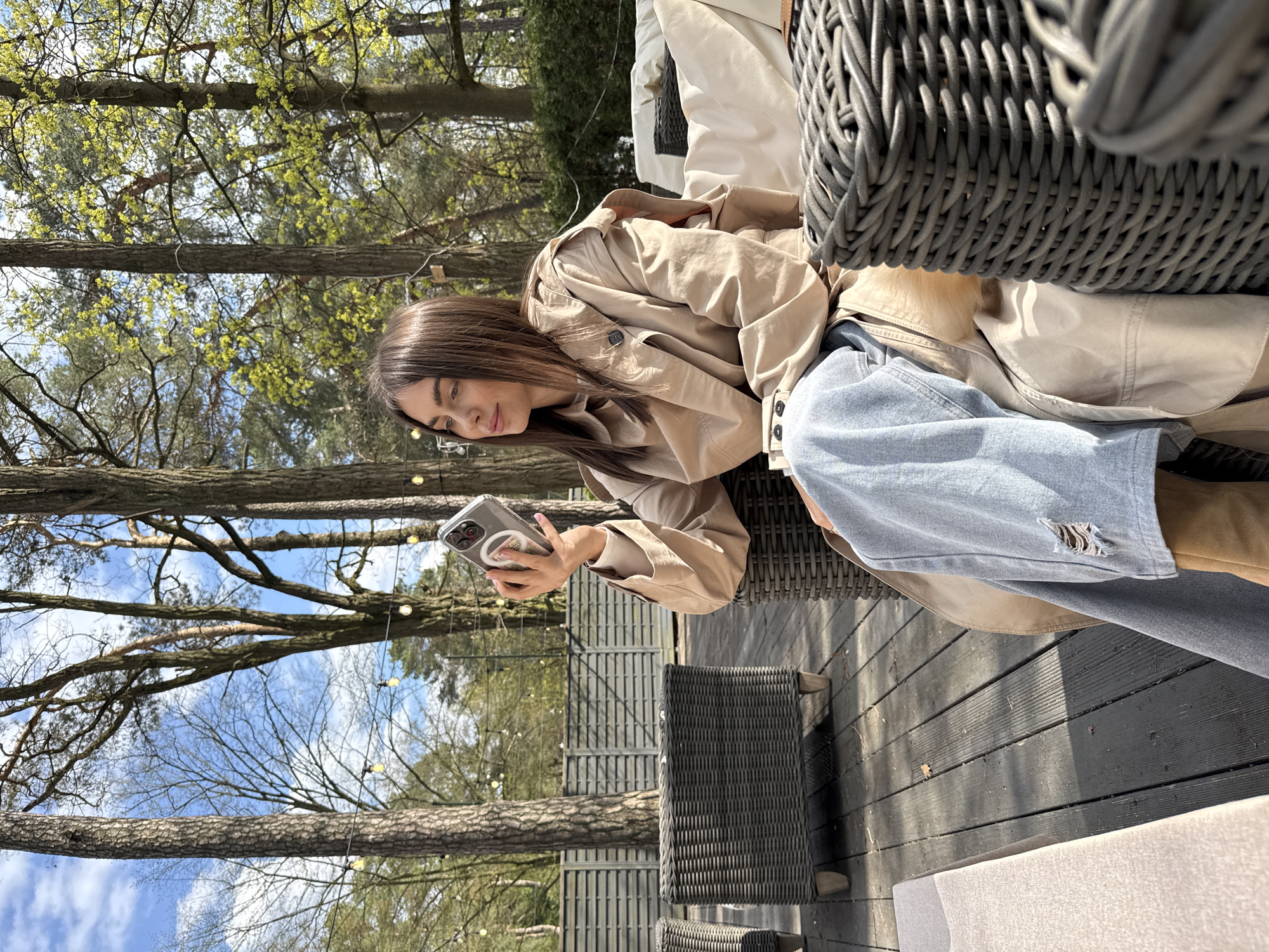 The image shows a woman seated outdoors, likely on a patio or deck with wicker furniture, surrounded by trees. She is looking at her cell phone, which she holds in her hands. She is wearing a trench coat and jeans. The location appears to be Konstancin-Jeziorna, Poland, based on visual elements such as the architecture and landscape.