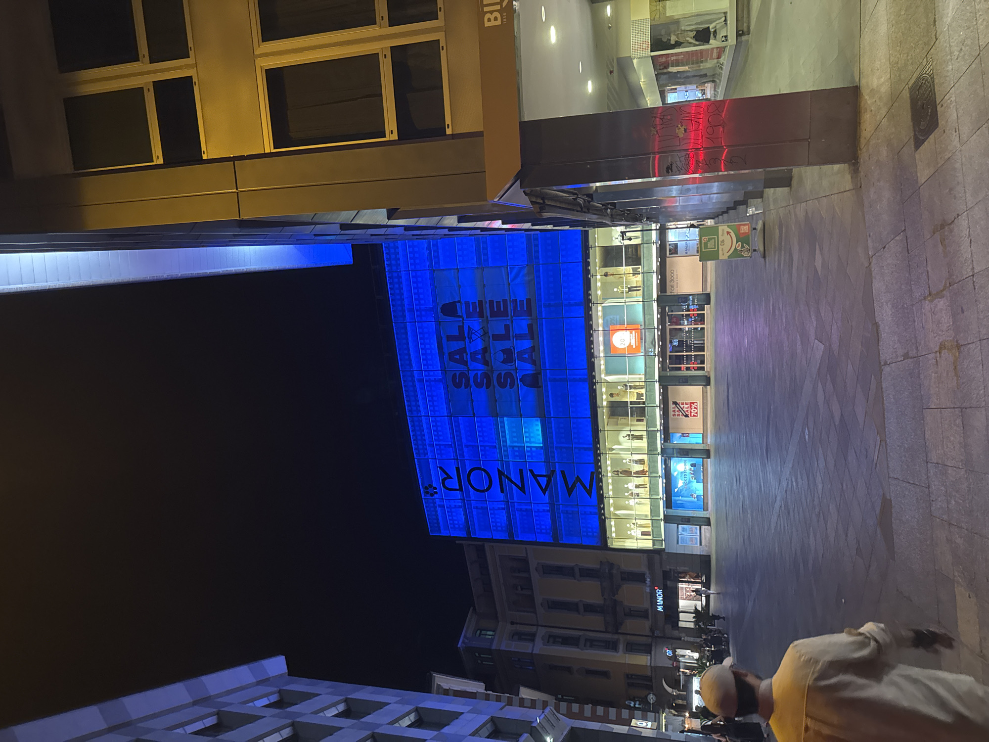 In the city of Lugano, Switzerland, at night, a broad, empty plaza is illuminated by the vibrant blue facade of the Manor department store. The store's upper floors are covered in a massive, backlit display of the word "MANOR" in large white letters, with "SALE" repeated in red text. Below this, the store's ground floor is visible through large glass windows, showcasing mannequins dressed in various outfits, suggesting a retail environment. The plaza's paving stones reflect the blue light. To the right of the Manor building, a more modern structure with yellow-tinted lighting and metal framing stands. A lone individual, wearing a light-colored shirt and a cap, walks away from the camera across the plaza, their back to the viewer. The overall atmosphere is one of a quiet, modern urban space at night, with the prominent store acting as a central visual element.
