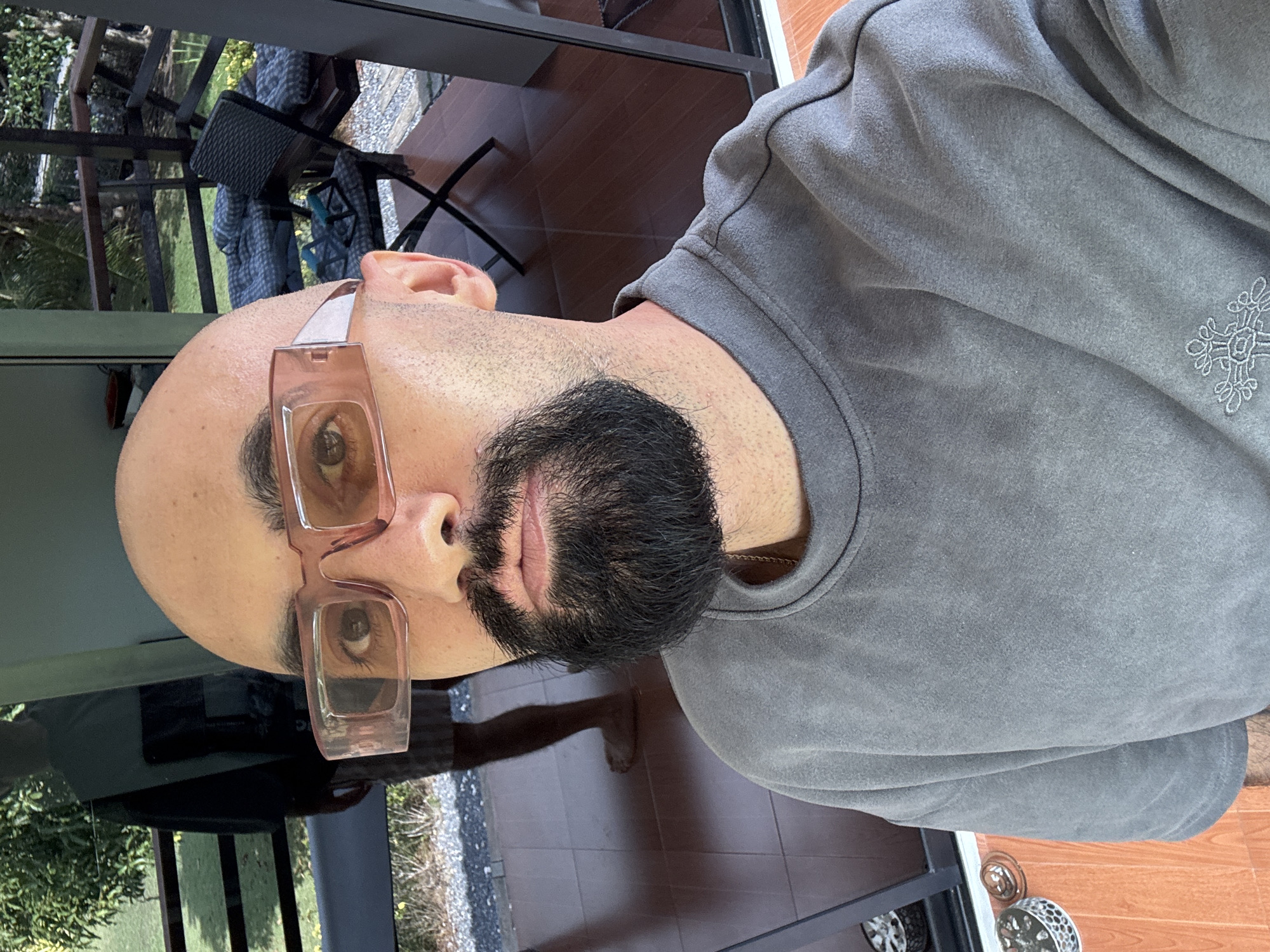 The image shows a close-up selfie of a bald man with a beard, wearing a gray sweatshirt and pink-tinted rectangular glasses. He is standing on a tiled balcony with a glass railing, overlooking a lush green outdoor area with trees and foliage. In the background, a reflection in the glass shows another person standing. A chair and some cushions are visible on the balcony. The overall setting appears to be a tropical location, potentially a resort or a private residence in Ko Samui District, Thailand, given the vegetation and outdoor living space.
