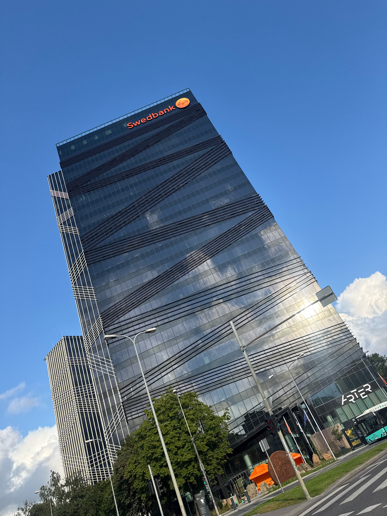 The image depicts the Swedbank headquarters building in Tallinn, Estonia. The modern skyscraper features a dark, glass facade with distinctive diagonal lines across its surface. The Swedbank logo is prominently displayed in orange at the top of the building. The building is surrounded by greenery and a street with traffic. A bus is visible on the right side of the image, and streetlights and a traffic signal are also present. The sky is a clear blue with some scattered clouds.