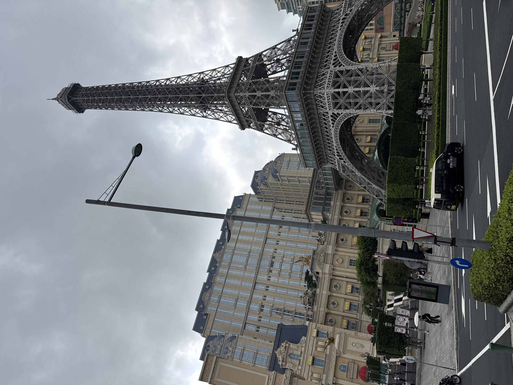 The image depicts a partial view of a replica of the Eiffel Tower, situated next to a large, ornate hotel building in the city of Unknown, Macao. The tower is grey in color and extends upwards into a cloudy sky. Below the tower, a street is visible with white lane markings, a black vehicle, and pedestrians. There's also a traffic light, signage, and a row of trimmed hedges. A tall street light stands in the foreground on the left.