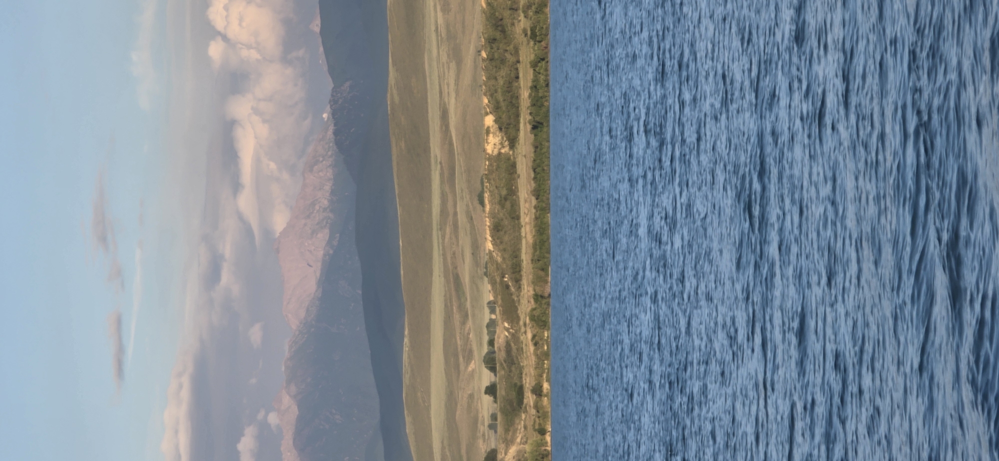 The image depicts a serene natural landscape. The foreground is dominated by a vast expanse of blue water, with gentle ripples disturbing its surface. Beyond the water, a treeline borders the shore, giving way to rolling hills covered in dry, golden-brown grass. Further in the distance, layered mountain ranges rise, their peaks softly illuminated by the sky. The sky itself is a pale blue, with scattered clouds, some appearing wispy and thin, others more substantial and tinged with pink. The overall impression is one of tranquility and natural beauty.